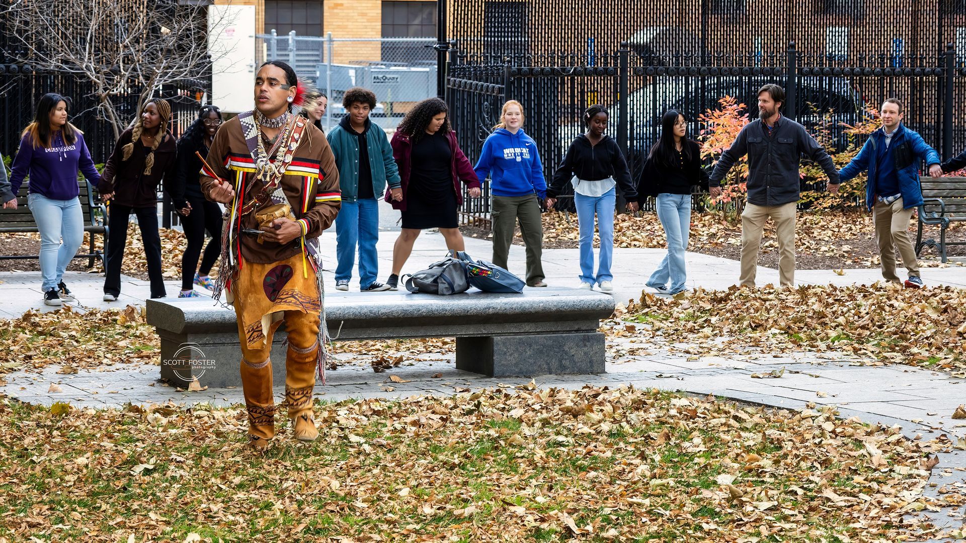 A Native American man performs a ceremony in a park, while a diverse group holds hands on a bench.