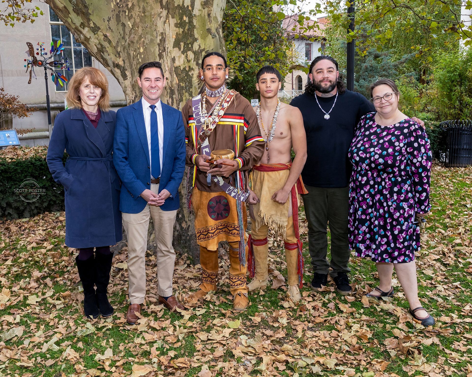 Group of six people posing outside, two in traditional Native American attire, autumn leaves.