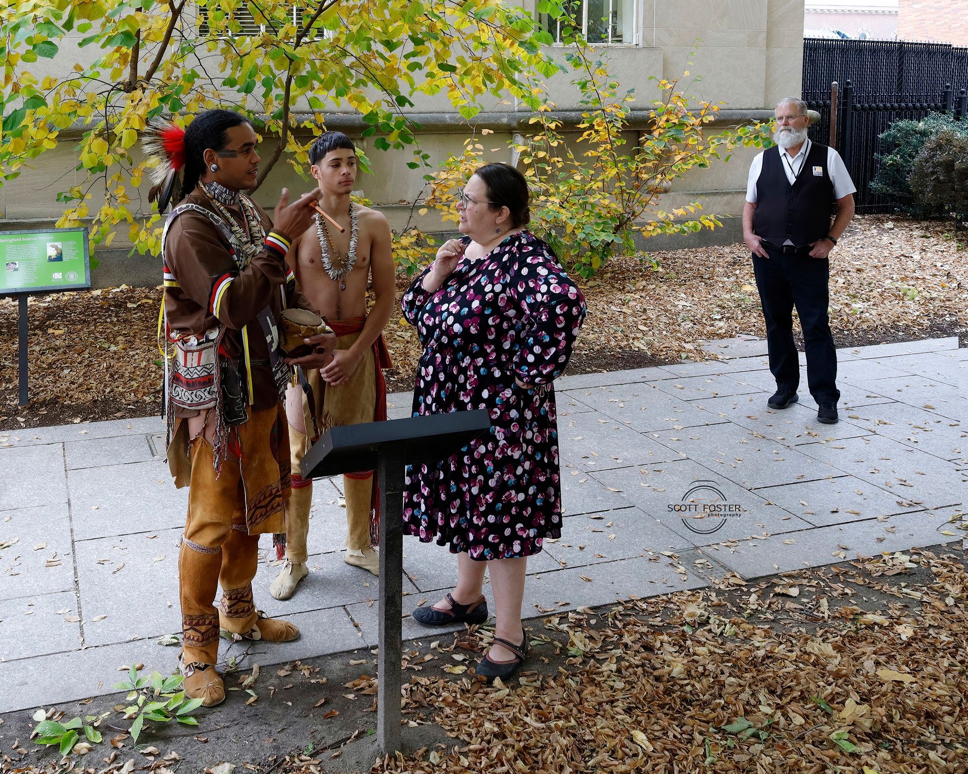 Two people in Native American attire speak with a woman near a sign; a man stands nearby.