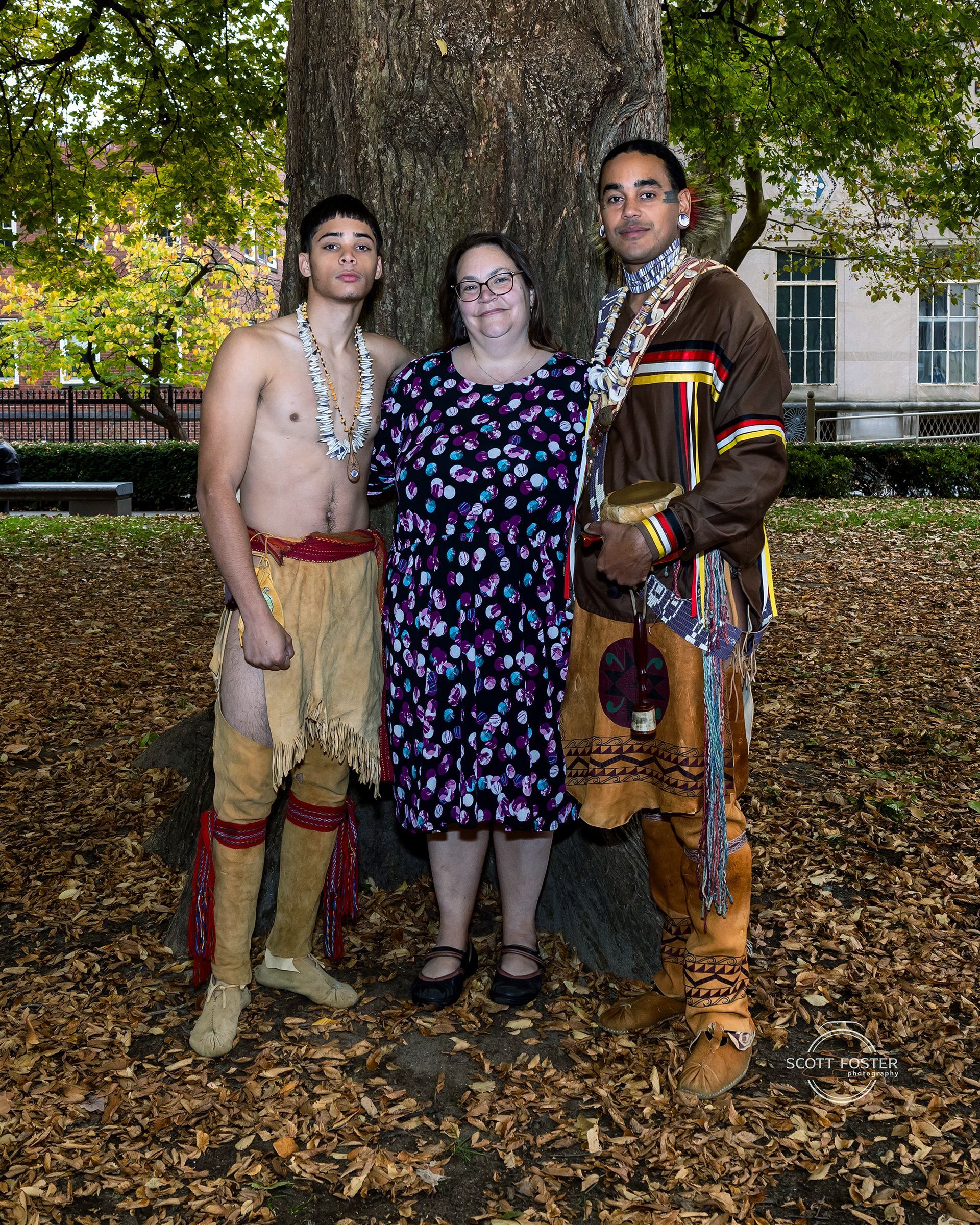 Woman stands between two people in Native American regalia, near a tree. Fall foliage surrounds them.
