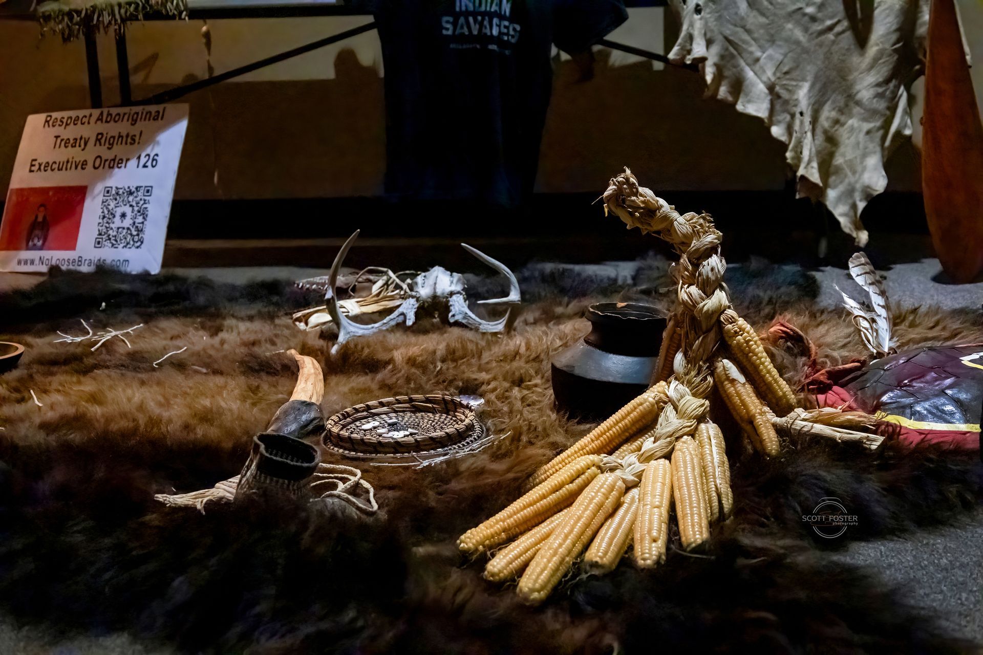 Display of Native American artifacts: corn, antlers, furs, a pot, and a sign.