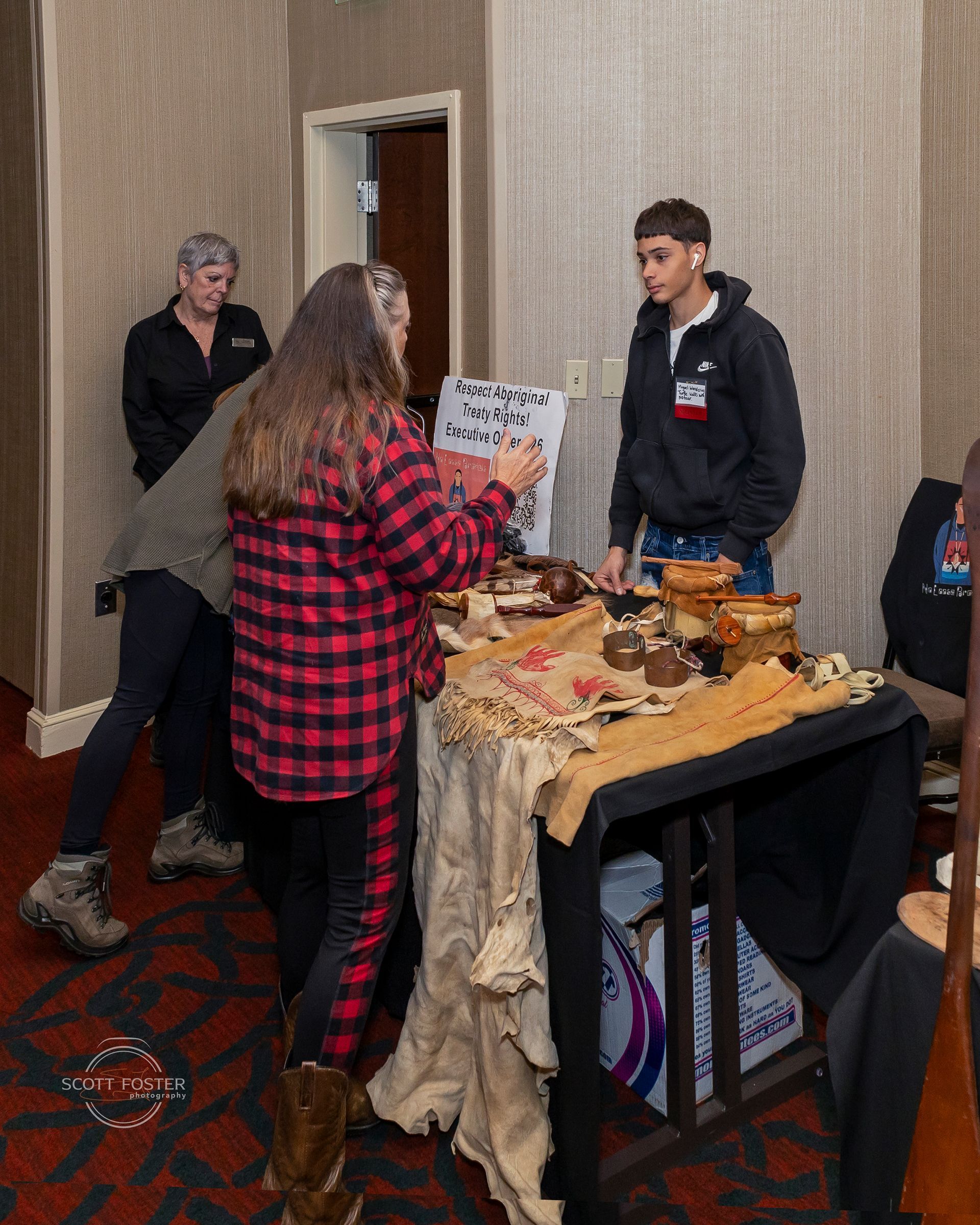 People at a booth with craft items. Woman in red plaid shirt points at display. Man in black hoodie stands by.