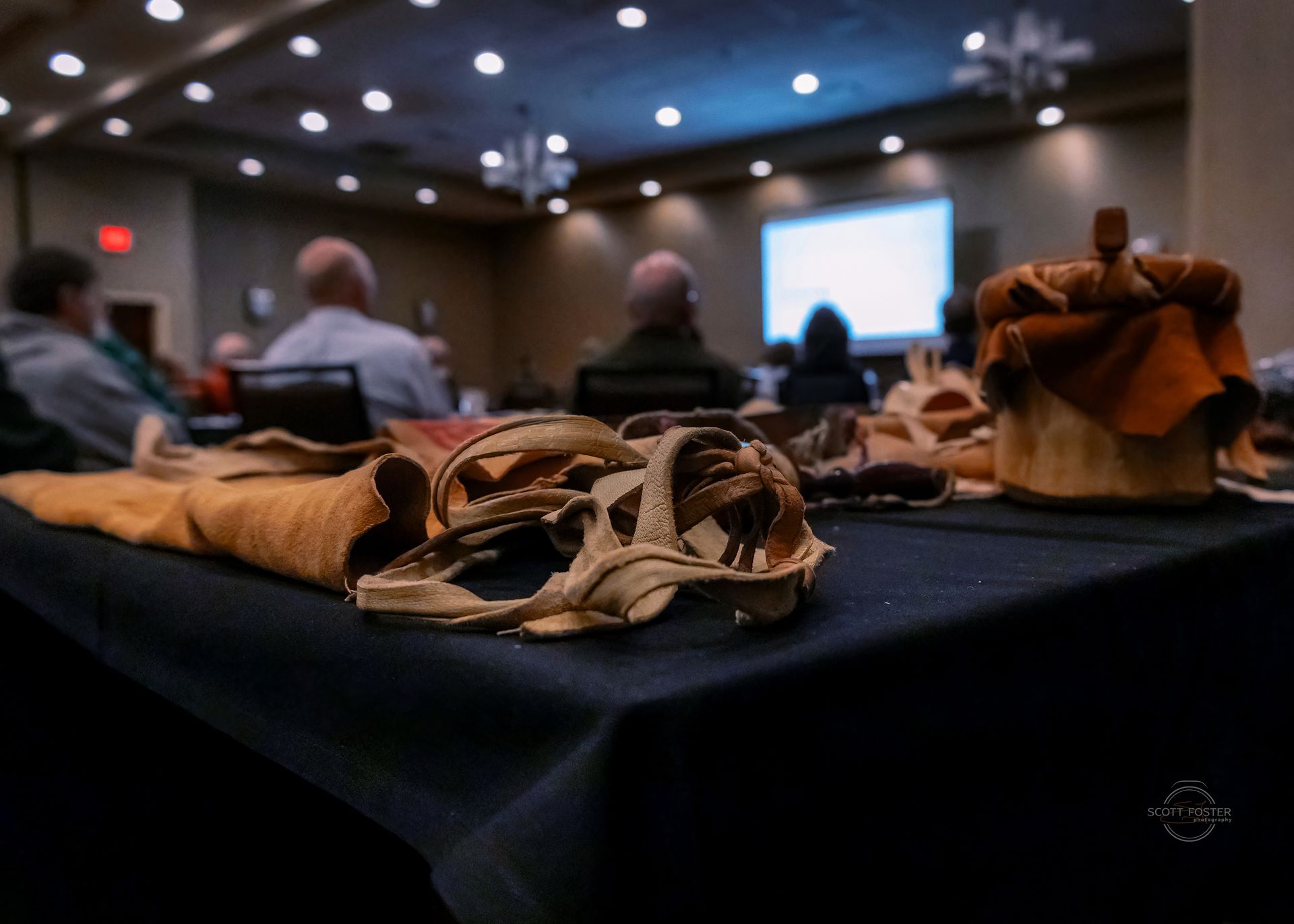 Leather crafts on table, presentation in background. Conference room with blurred audience, overhead lighting.