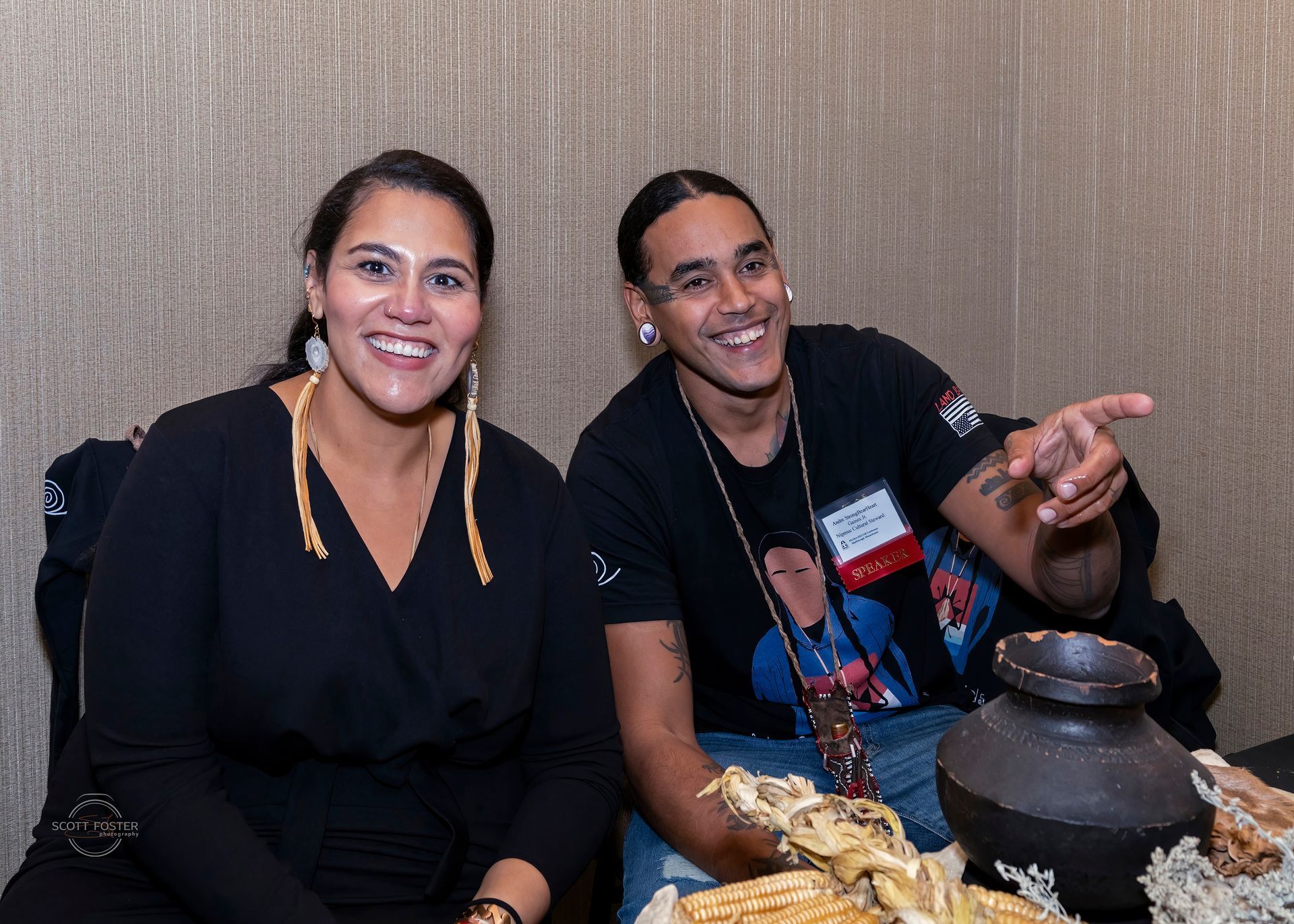 Two people smiling, one points. Seated next to food display in a room with textured wall.