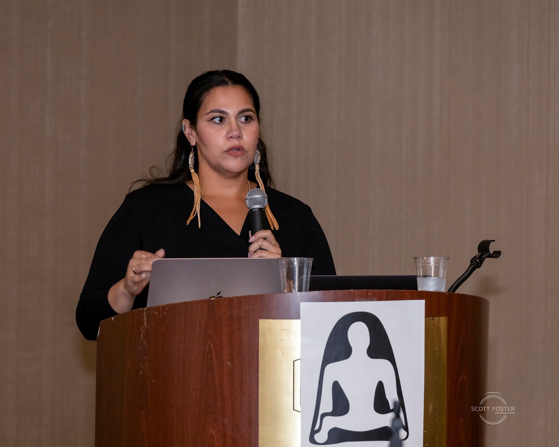 Woman speaking at podium, holding microphone, laptop visible. Artwork on podium. Indoor setting.