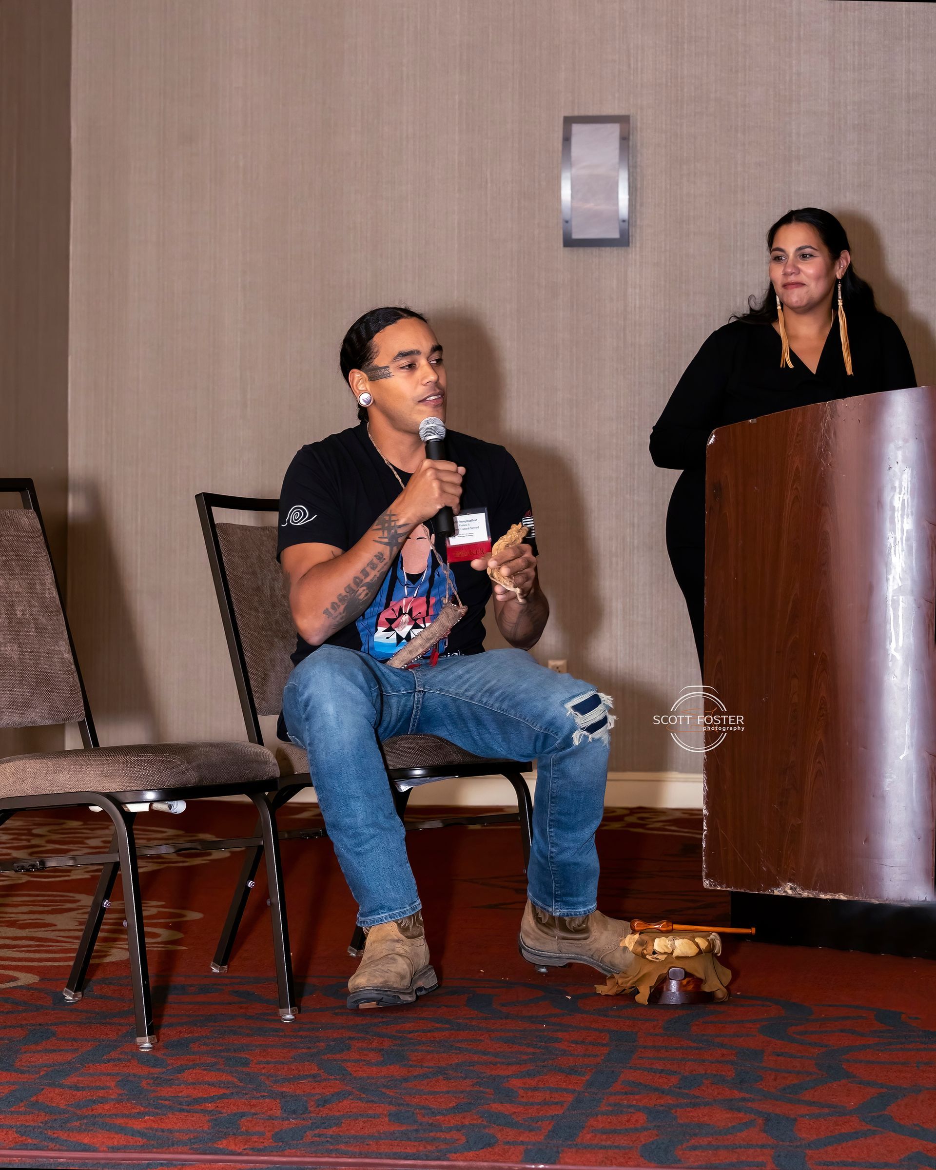 Man speaking into a microphone, sitting on a chair, another person stands by a podium.