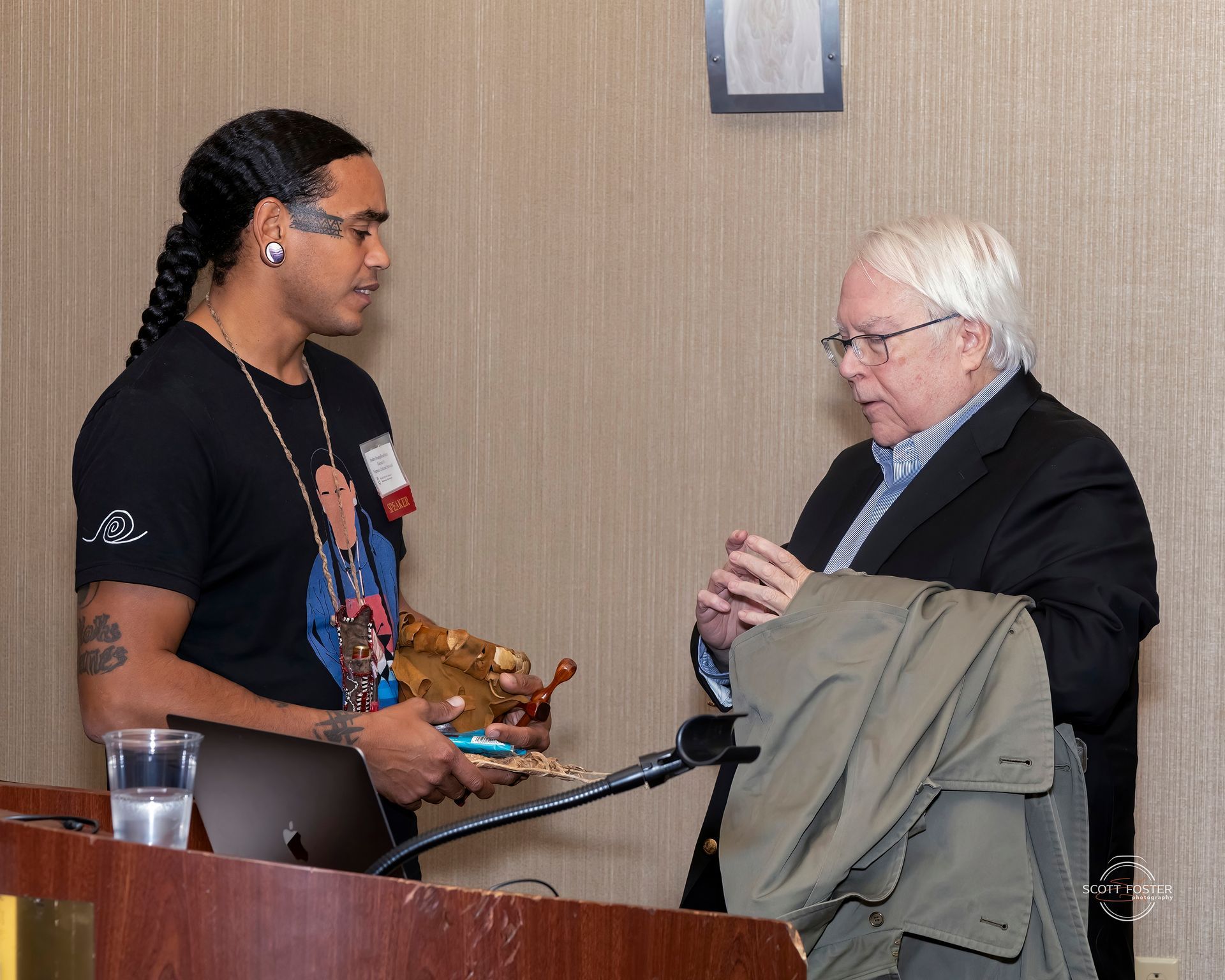 Man with long hair and tattoos presents a wooden object to an older man in a suit, near a podium.