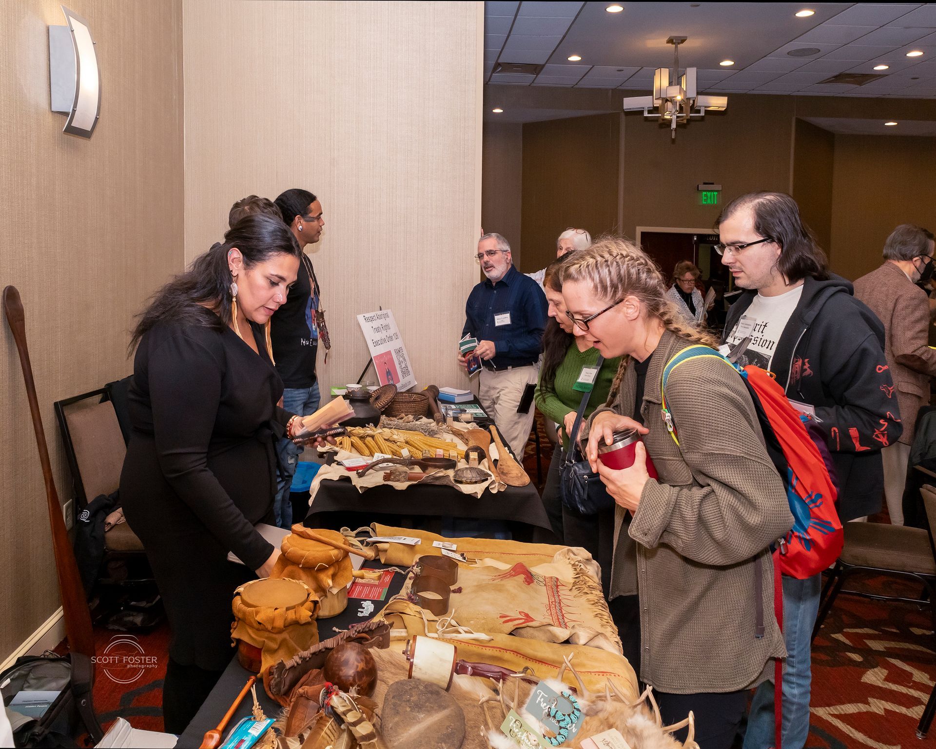People examining a display table with artifacts at an indoor event; a woman gestures.