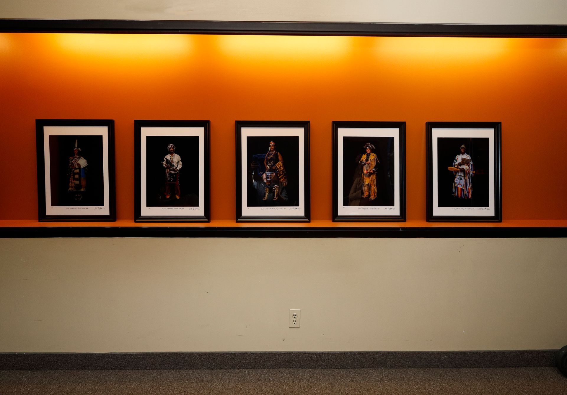 Five framed portraits on a shelf against an orange wall. The wall has a recessed light and is above a beige wall.
