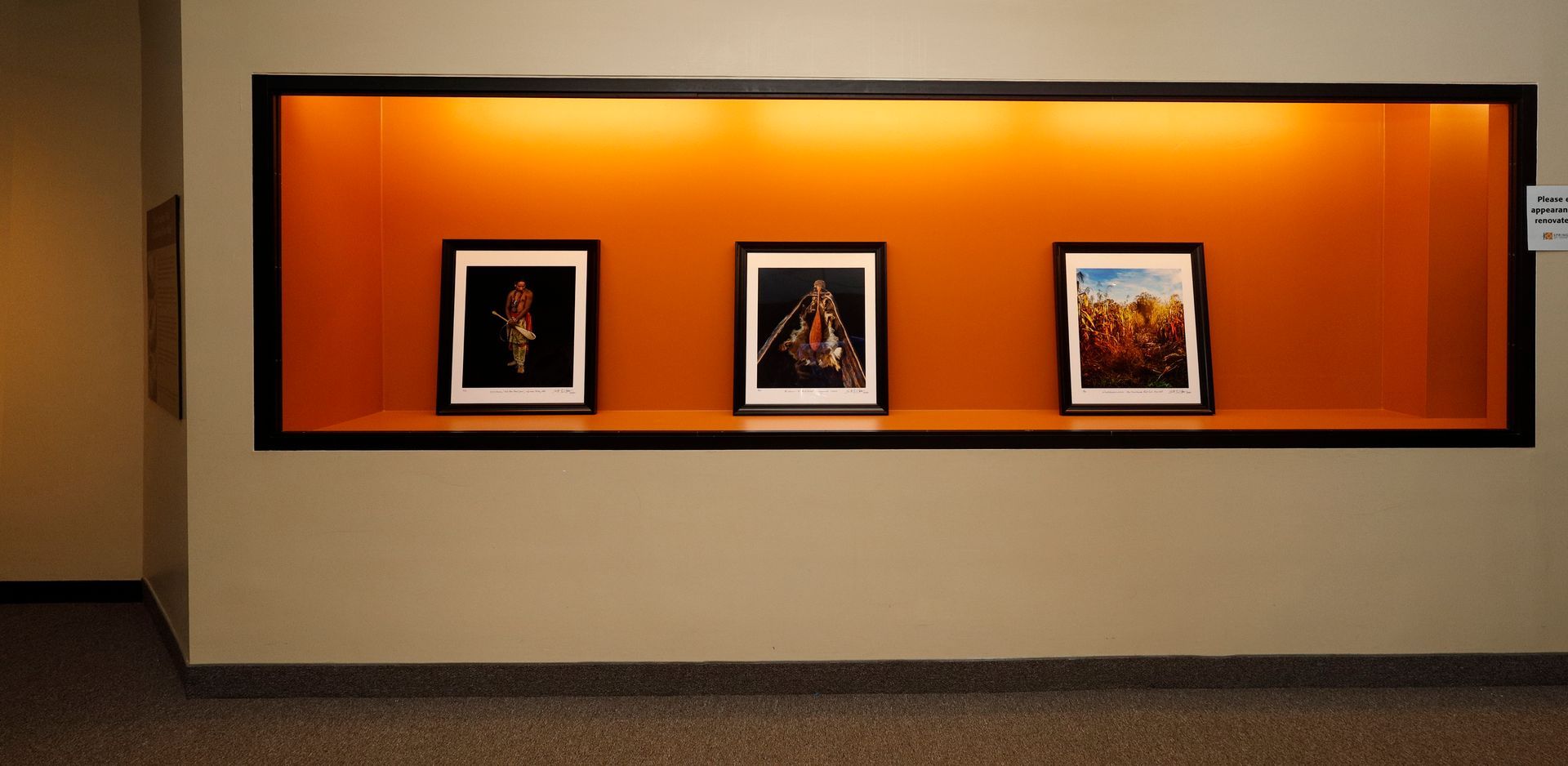 Three framed artworks displayed in an orange-lit niche on a beige wall.