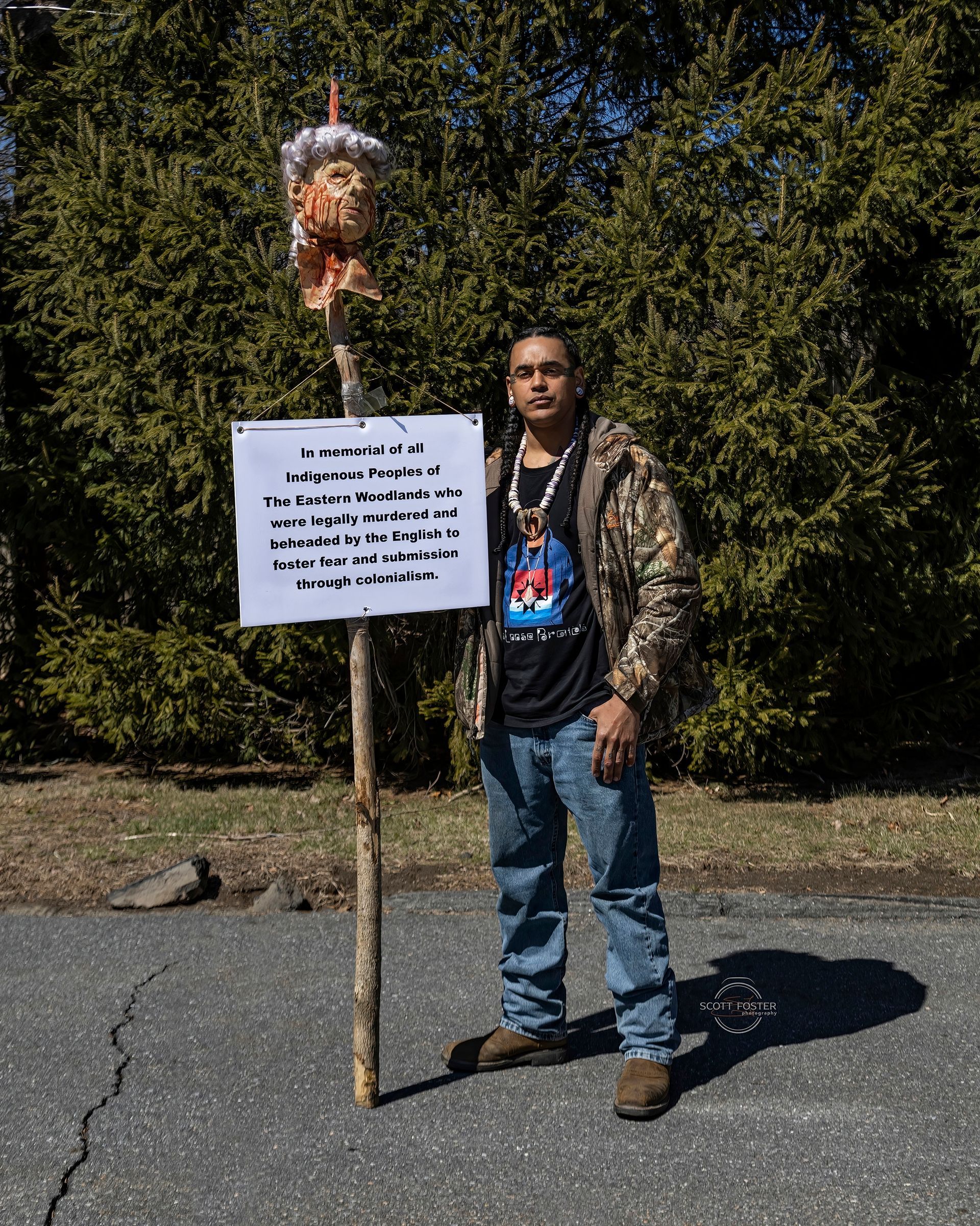 Man stands with sign and effigy on a stick. He wears jeans, camo jacket, and a shirt with a graphic.