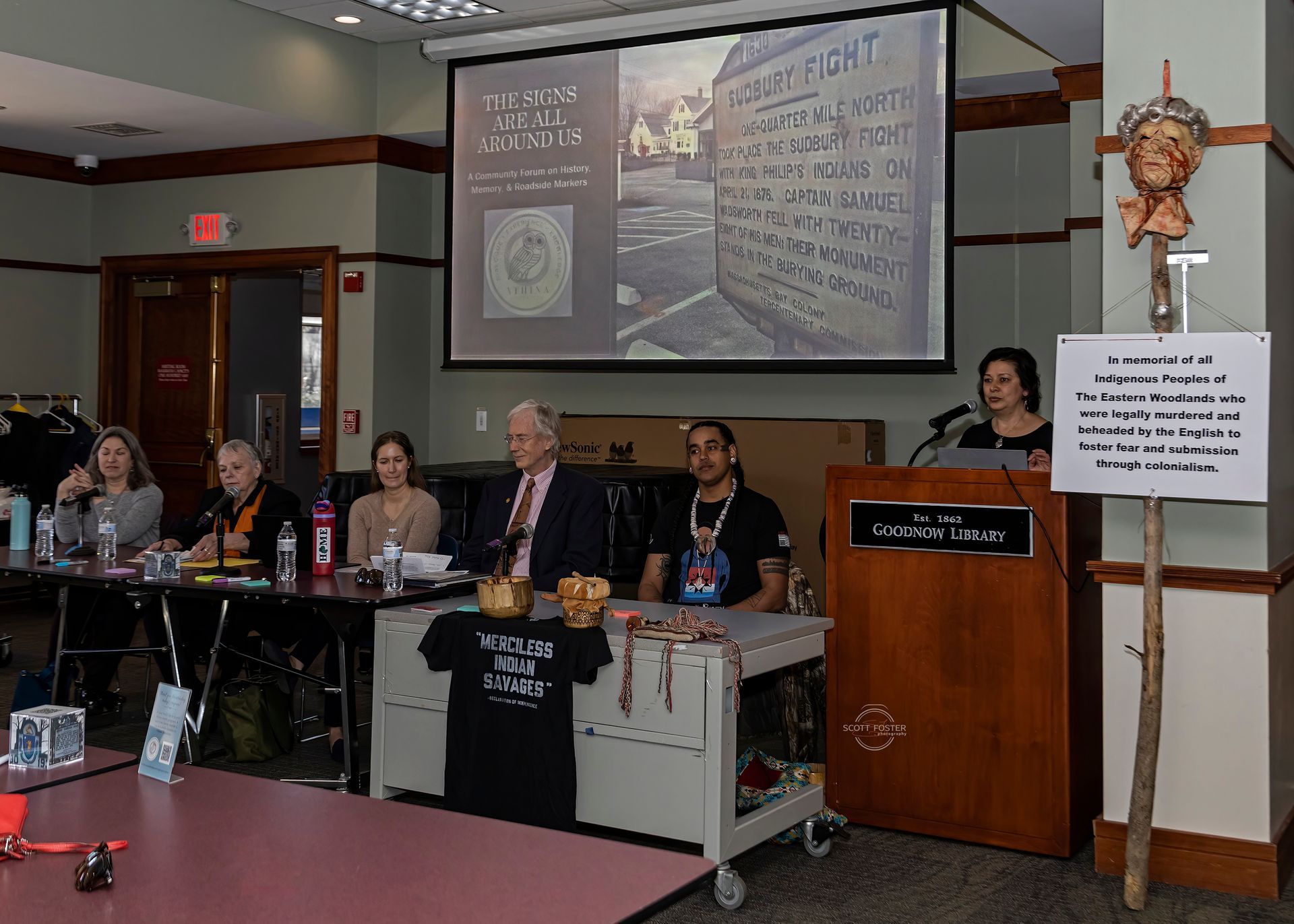 A presentation with speakers at a podium. A slideshow displays a monument. Artifacts and a sign are on display.