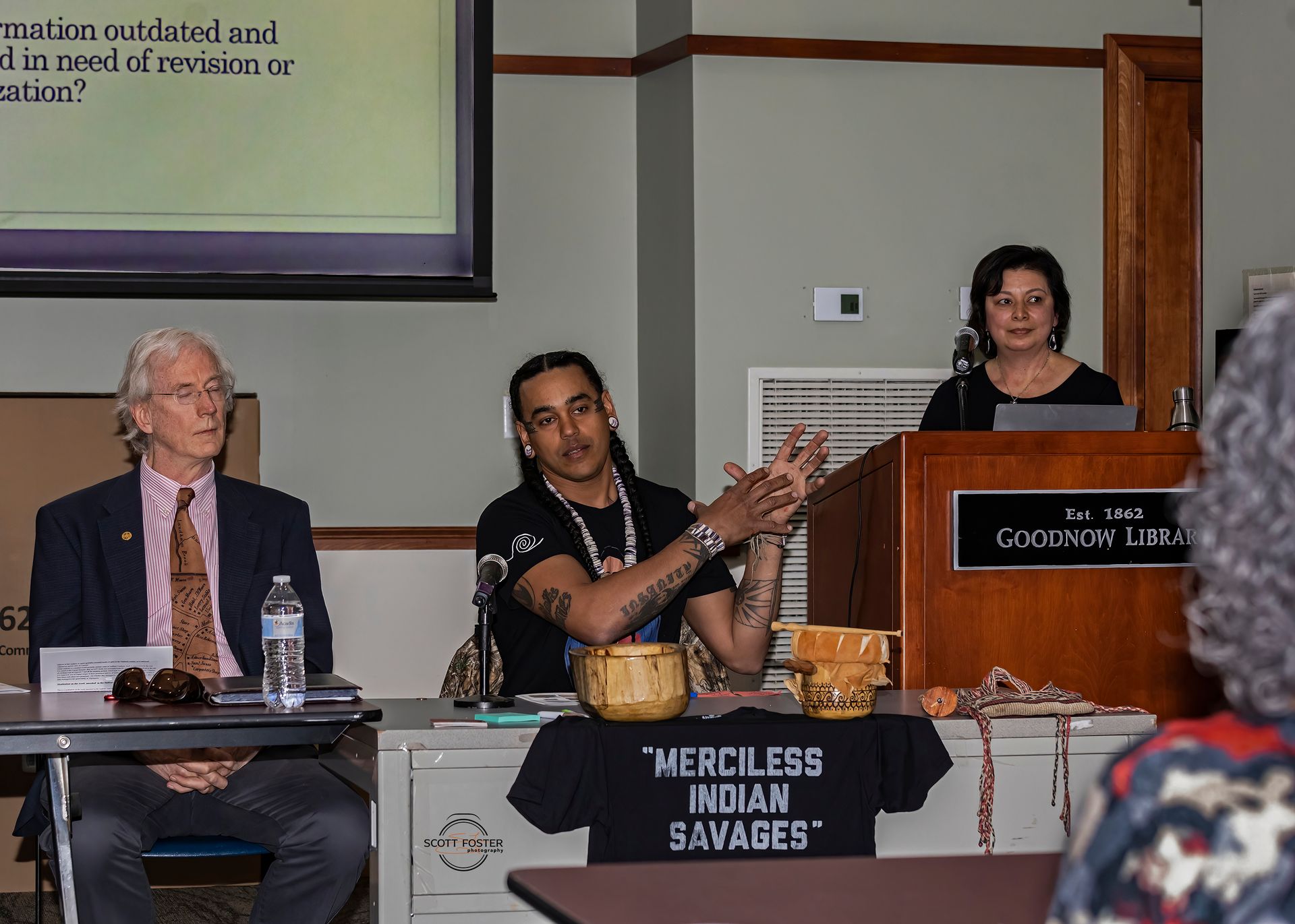 Three people at a podium during a presentation. Man gesturing, woman speaking. Sign says 