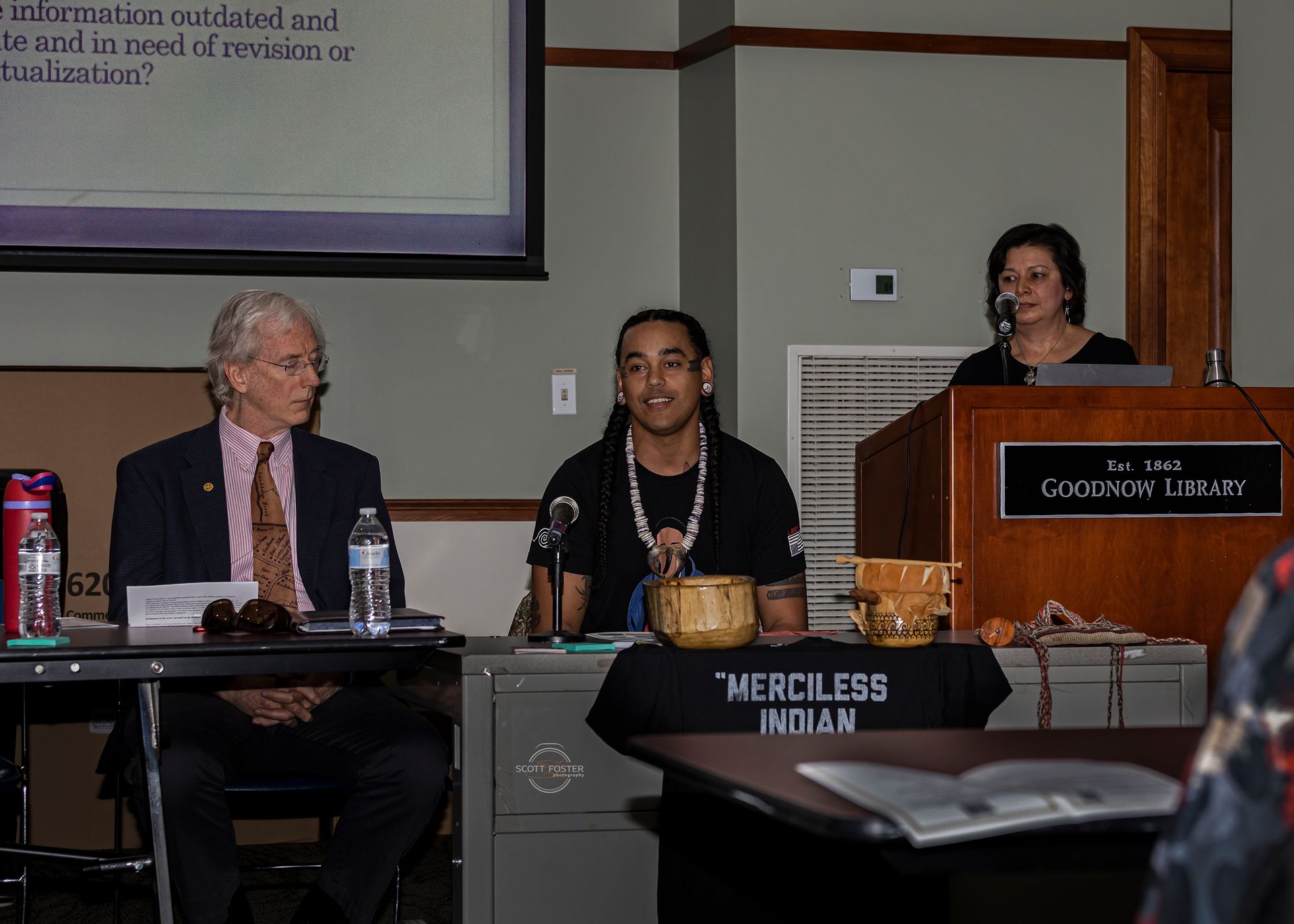 Three people at a podium in a library, one man speaks at a microphone, 