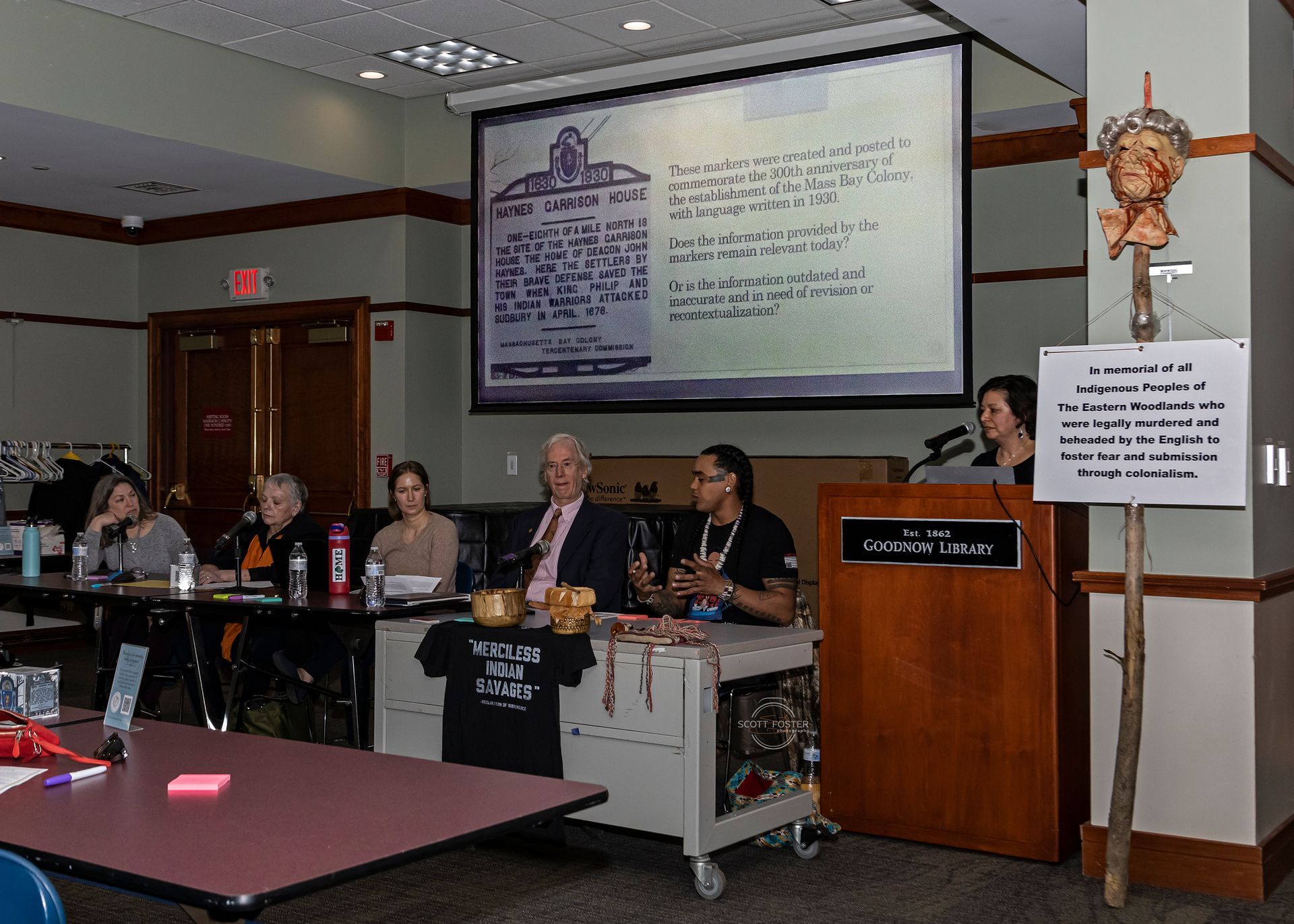 A panel discussion at a library. Speakers and audience at a table, with a projected document and a sign.