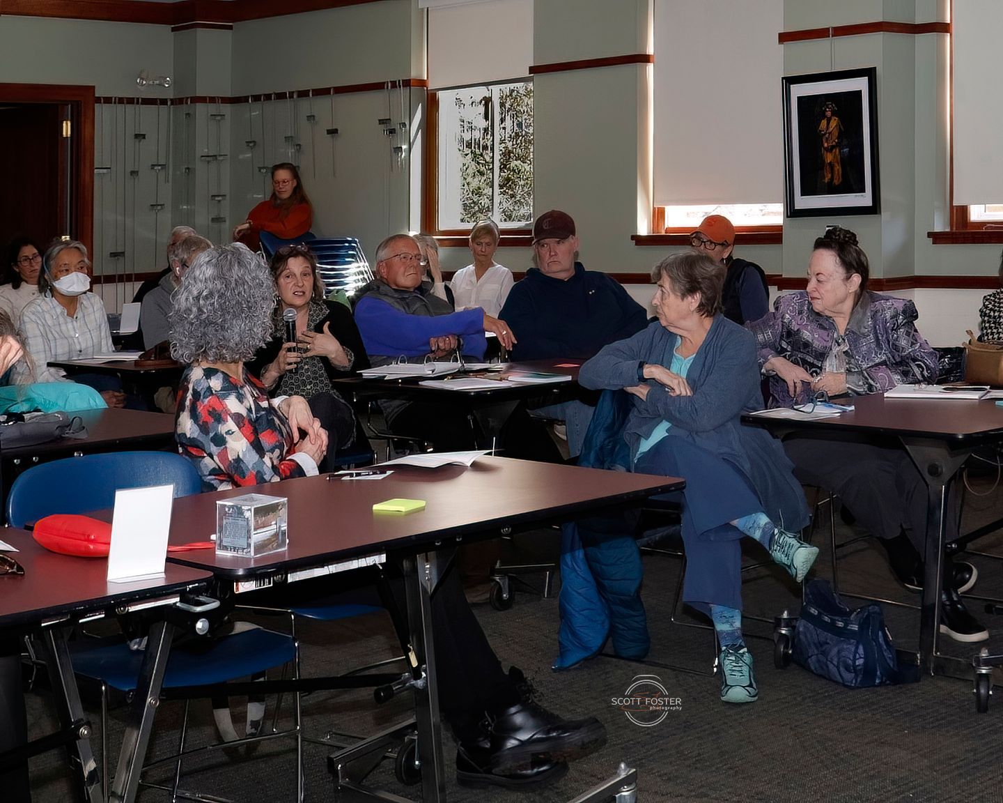 People seated at tables in a room, appearing to be in a meeting or class. Some are facing forward, others talking.