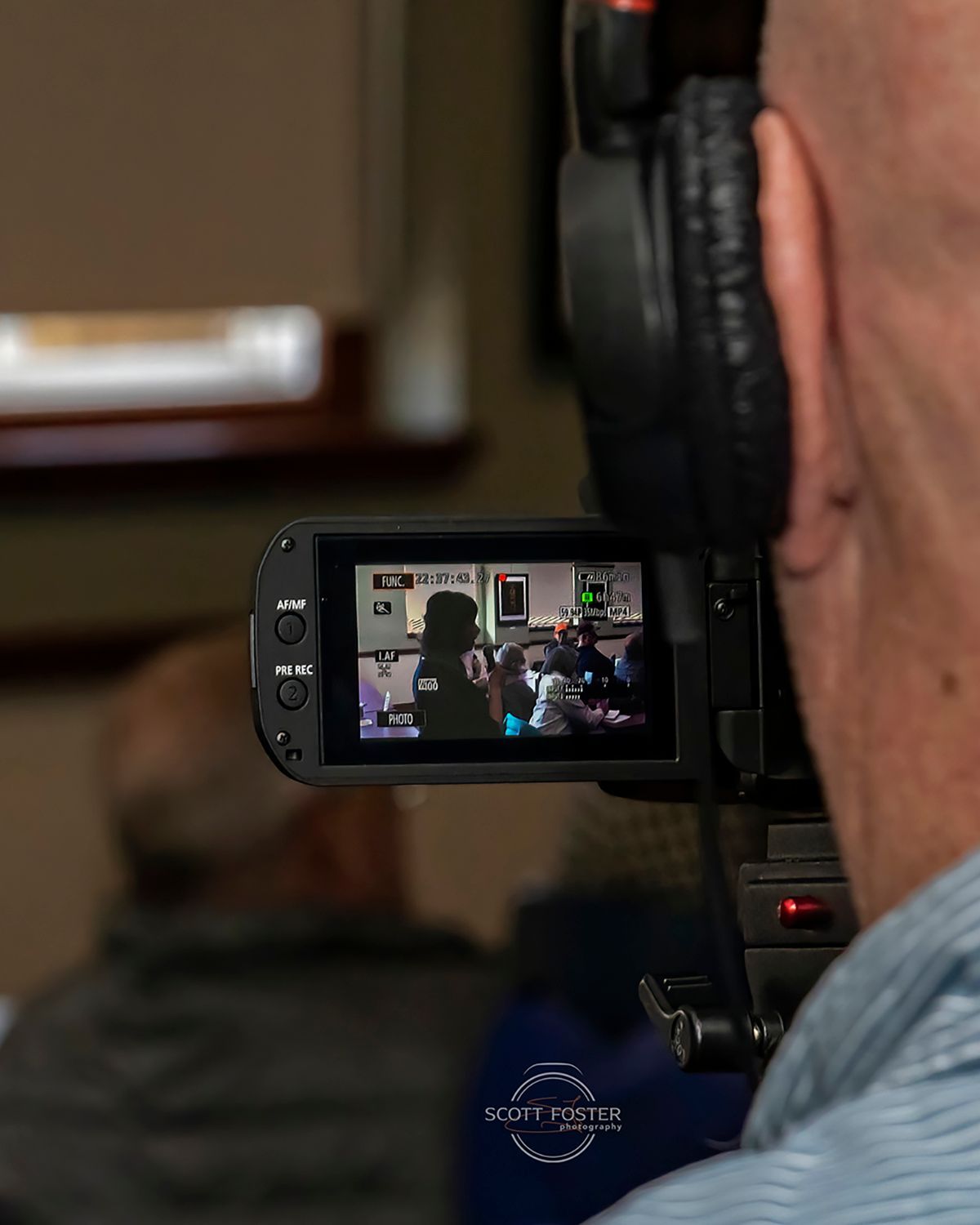 Person filming with a video camera; camera screen shows a seated person speaking.