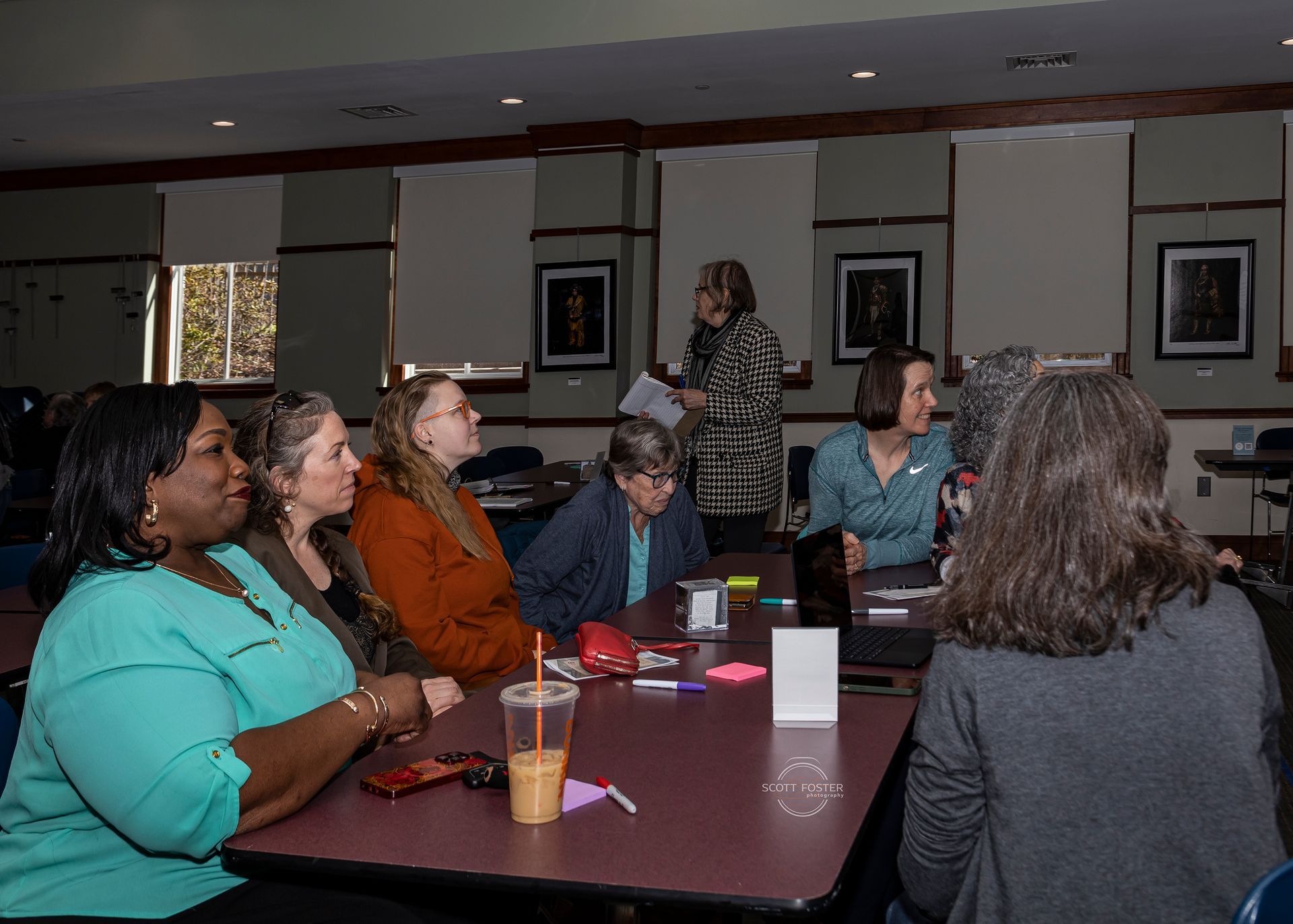 A group of people sitting around a table in a room, listening to a woman standing.