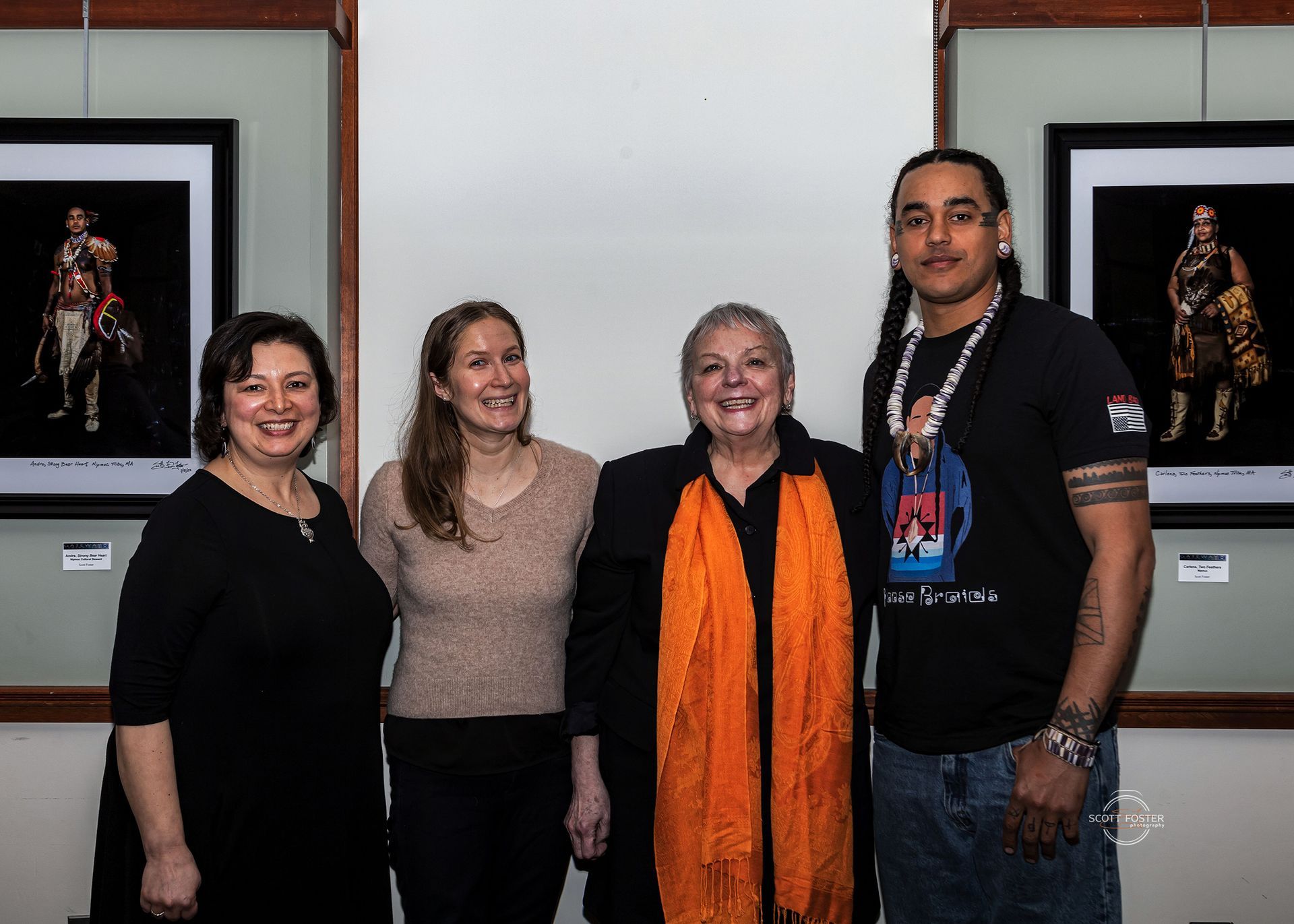 Four people stand together in front of framed artwork. One wears an orange scarf, and another has long hair.