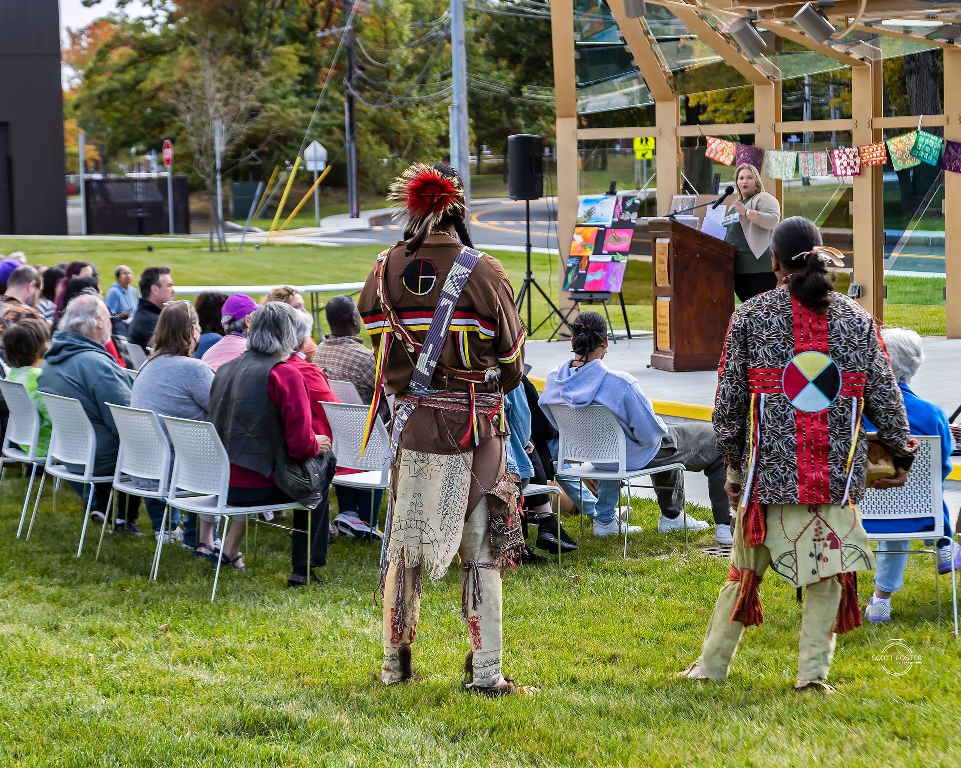 People at outdoor event; two in traditional clothing face crowd; speaker at podium, colorful decorations.