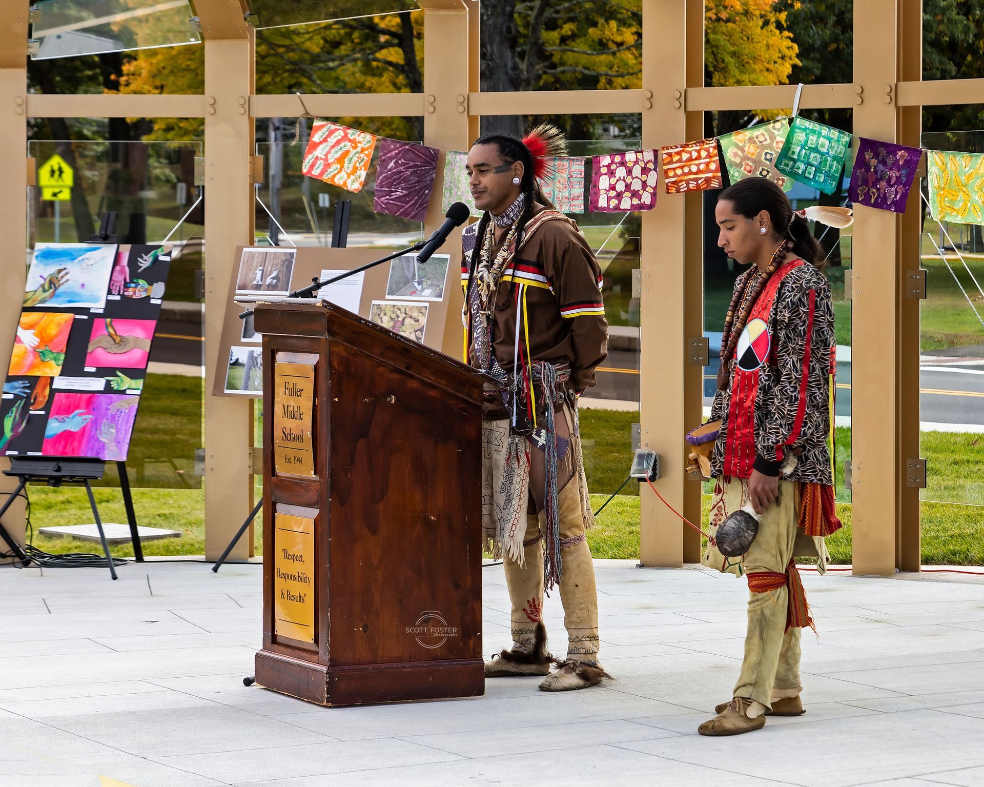 Two Indigenous men speaking at an outdoor event, one at a podium. Colorful artwork and banners are in the background.