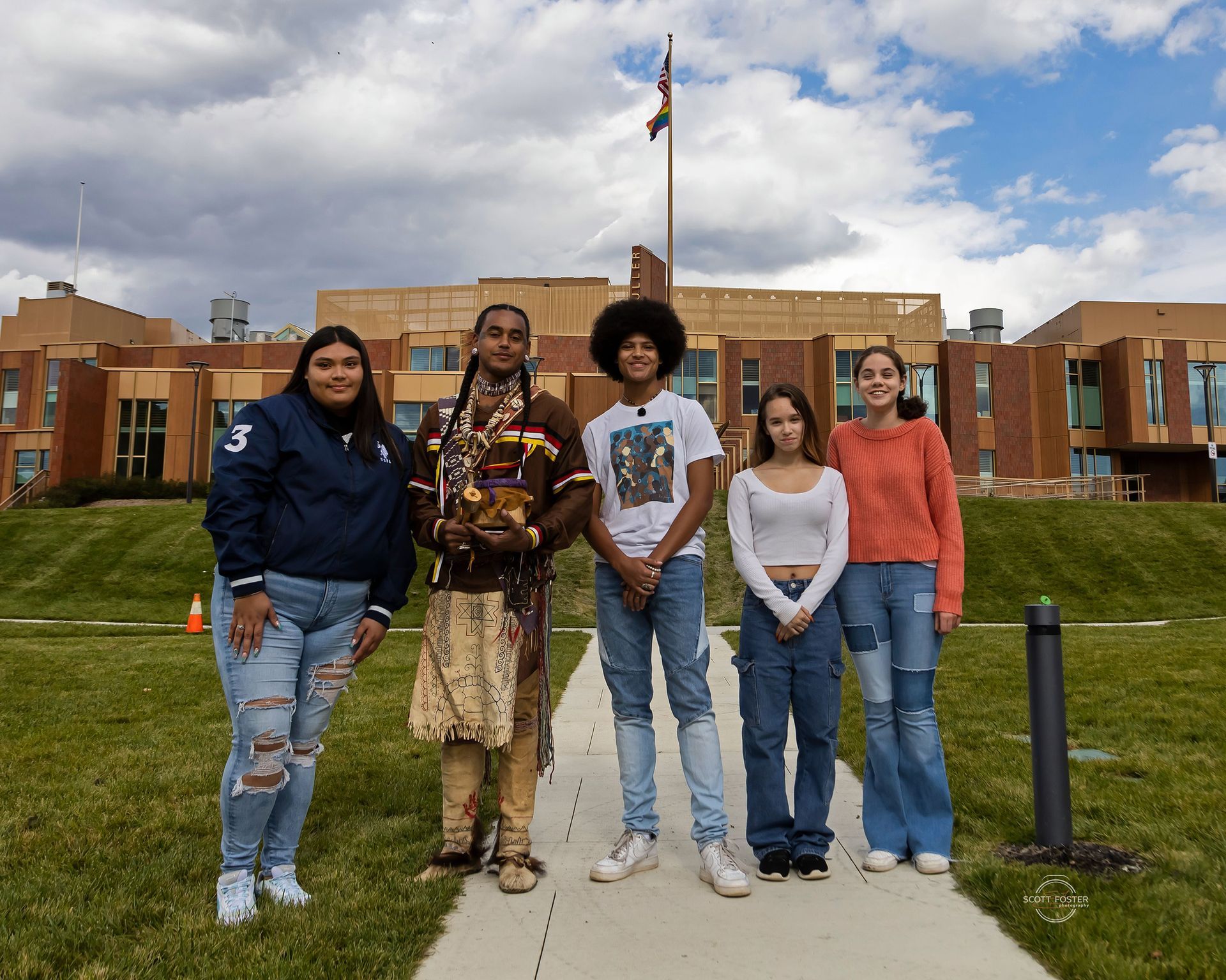 Five people stand on a walkway in front of a building. One person wears Indigenous regalia. 