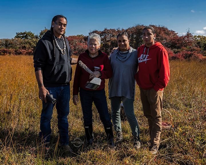 Four people in a field, one holding an award. Autumn setting with tall grass and blue sky.