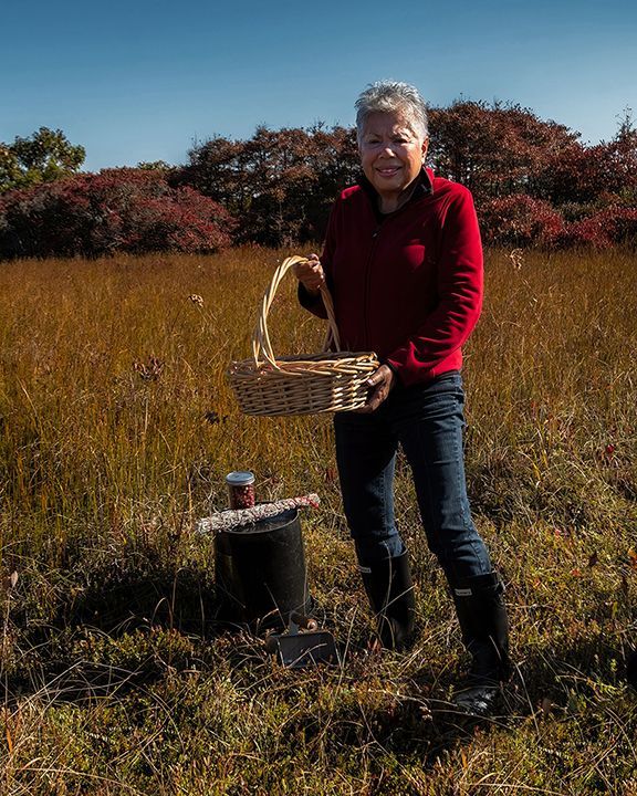Woman in red sweater and jeans holding a basket in a field, tall grass and fall foliage in the background.