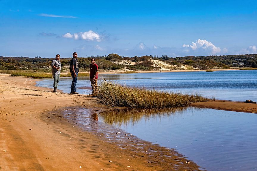 Three people standing on a sandy beach, looking out at a calm blue body of water on a sunny day.