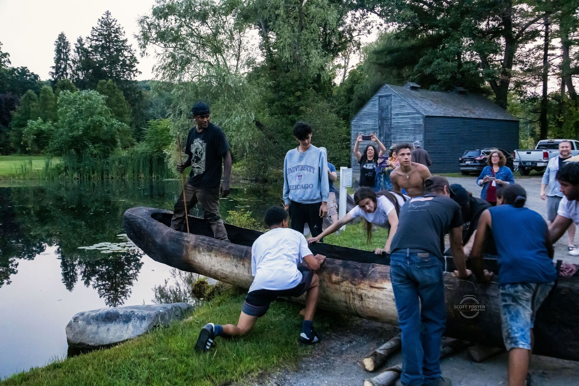 People pulling a large, charred log boat toward a lake's edge. Trees and a small building are in the background.