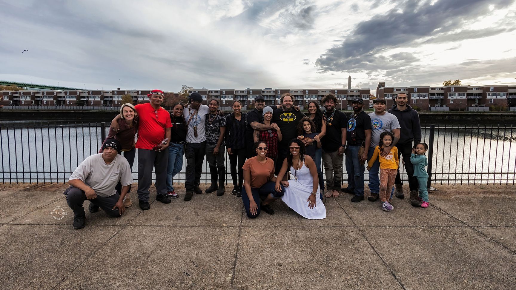 Group of people posing by a waterfront with townhouses in the background, under a cloudy sky.