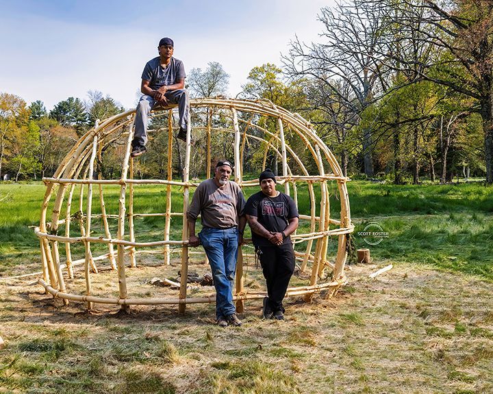 Three people pose near a wooden dome structure. Green grass and trees are in the background.