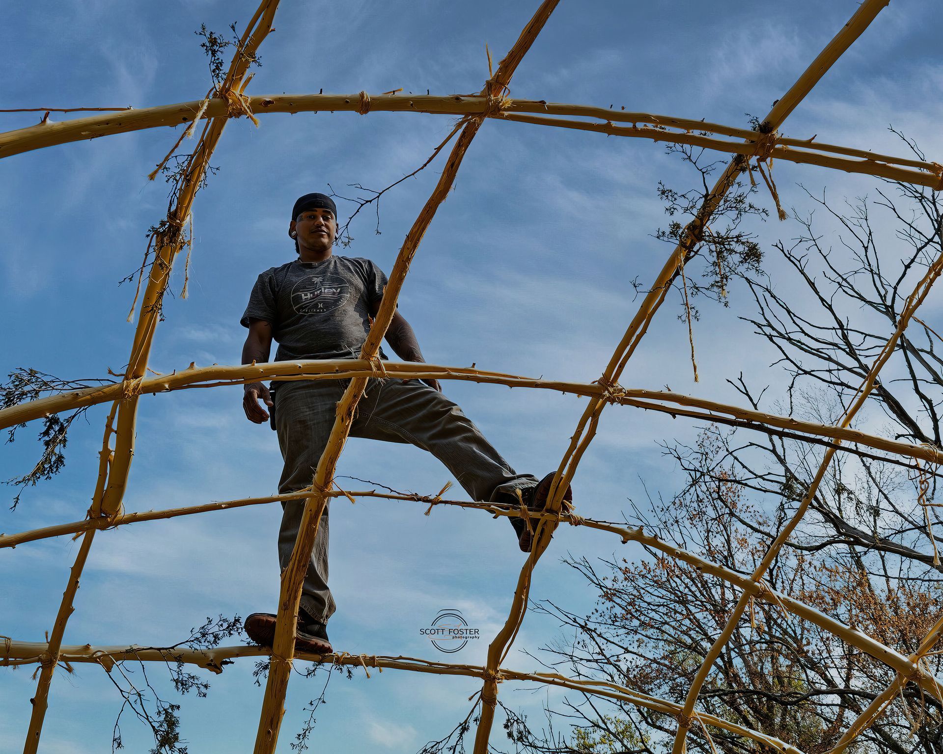 Man standing on a yellow, latticed structure against a blue sky. Bare trees are in the background.