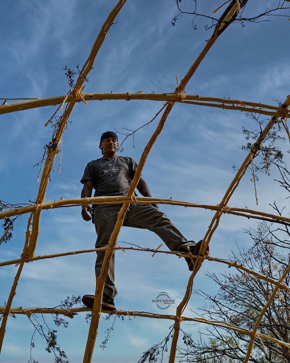 Man standing on a curved frame made of sticks against a blue sky.