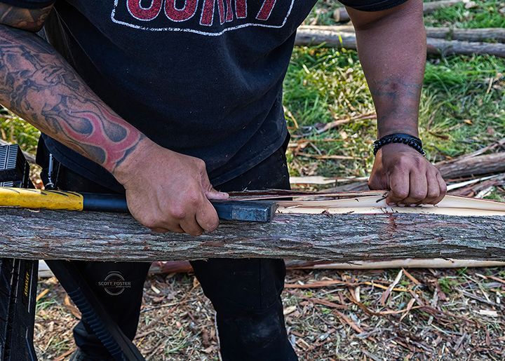 Person using a chisel to carve wood outdoors, wearing a black shirt and has arm tattoos.