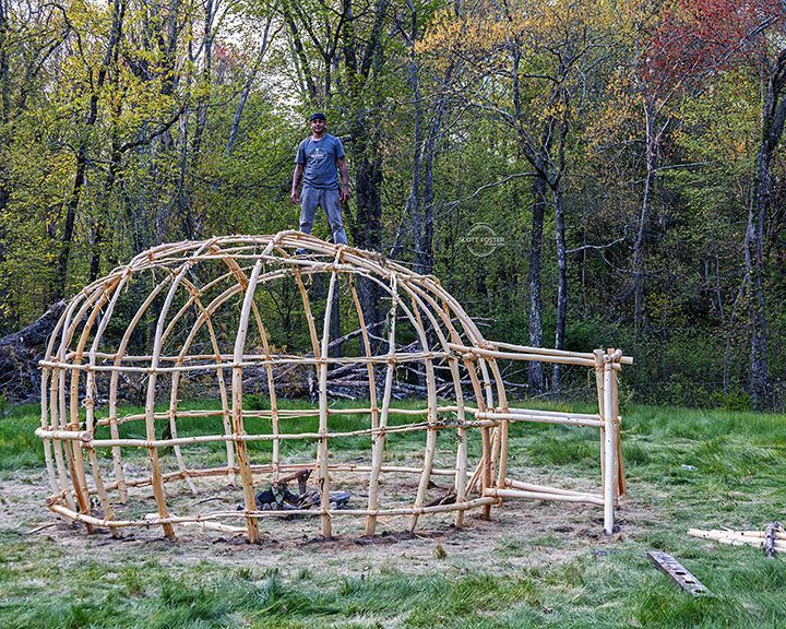 Man stands on a wooden frame structure, possibly a dwelling, in a grassy area surrounded by trees.