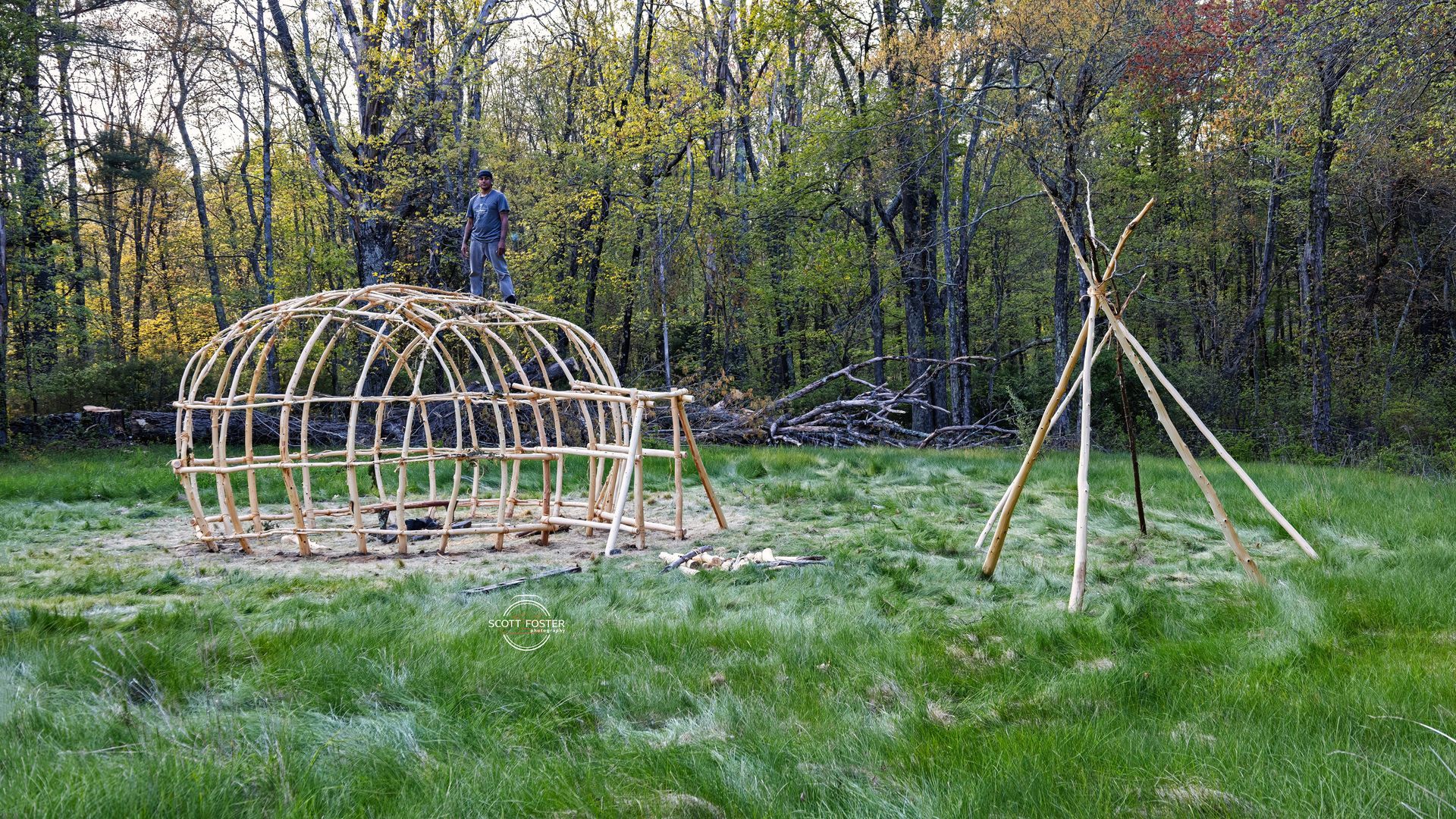 A wooden structure under construction in a grassy area with two people on top and a teepee-like frame.
