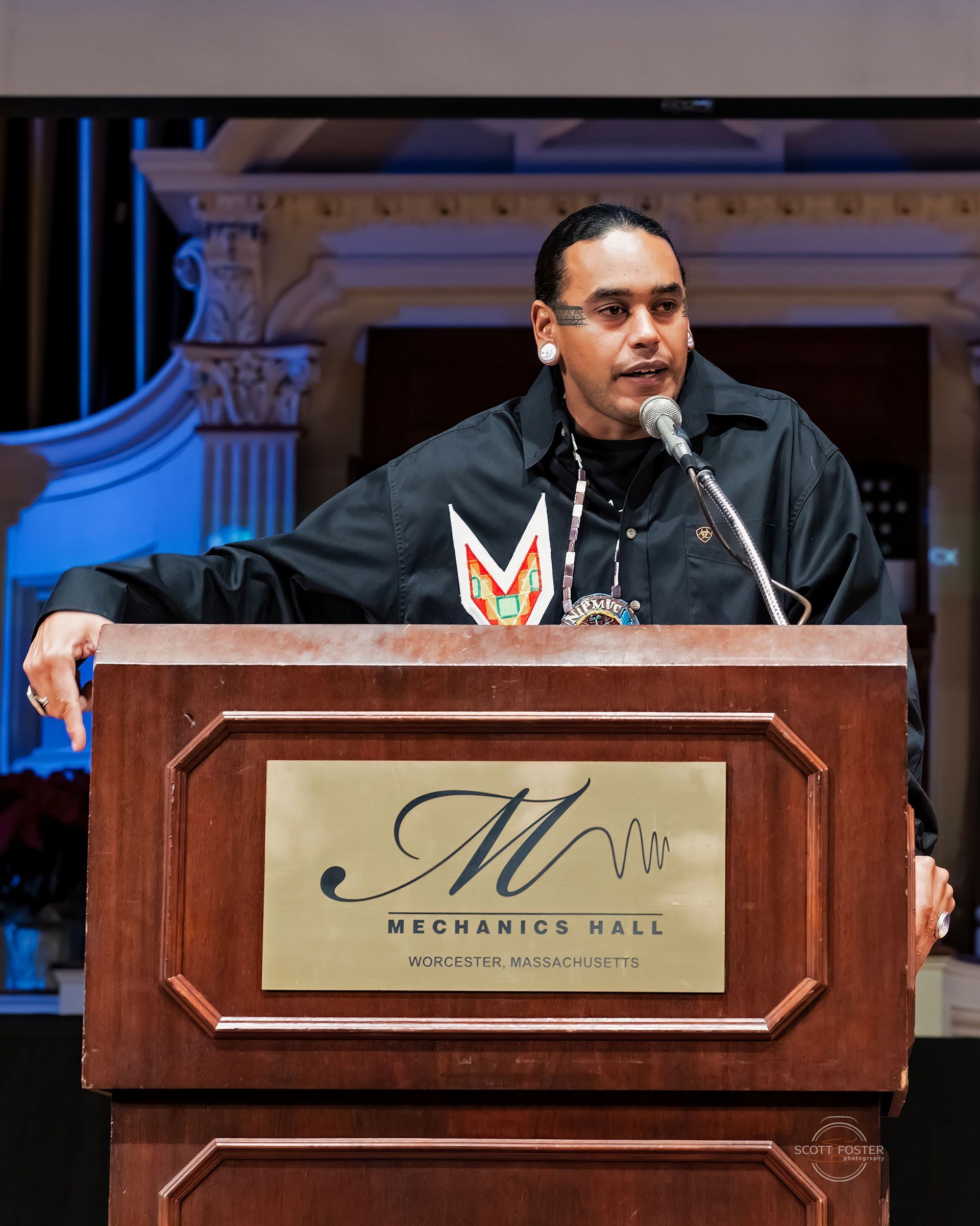 Man speaking at a podium with “MP CHARLES HALL” sign, wearing black shirt with Native American design.