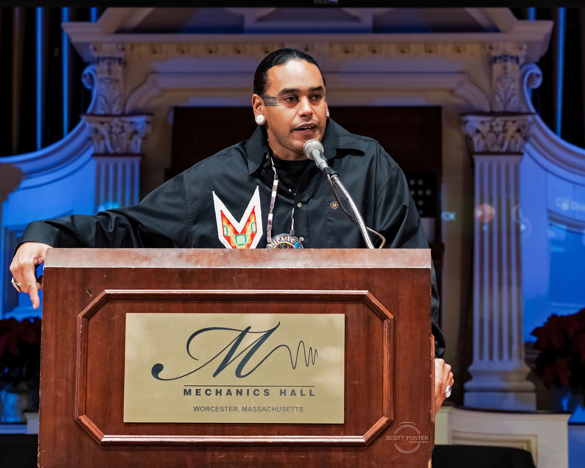 Man speaking at a podium in Mechanics Hall, wearing a black shirt with colorful design.