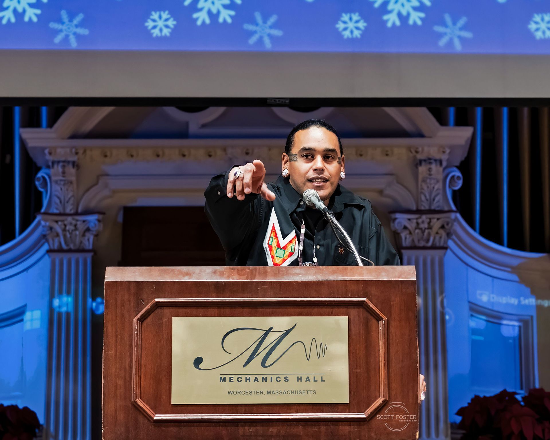 Man at a podium with microphone, gesturing. He is in front of an ornate backdrop with snowflakes.