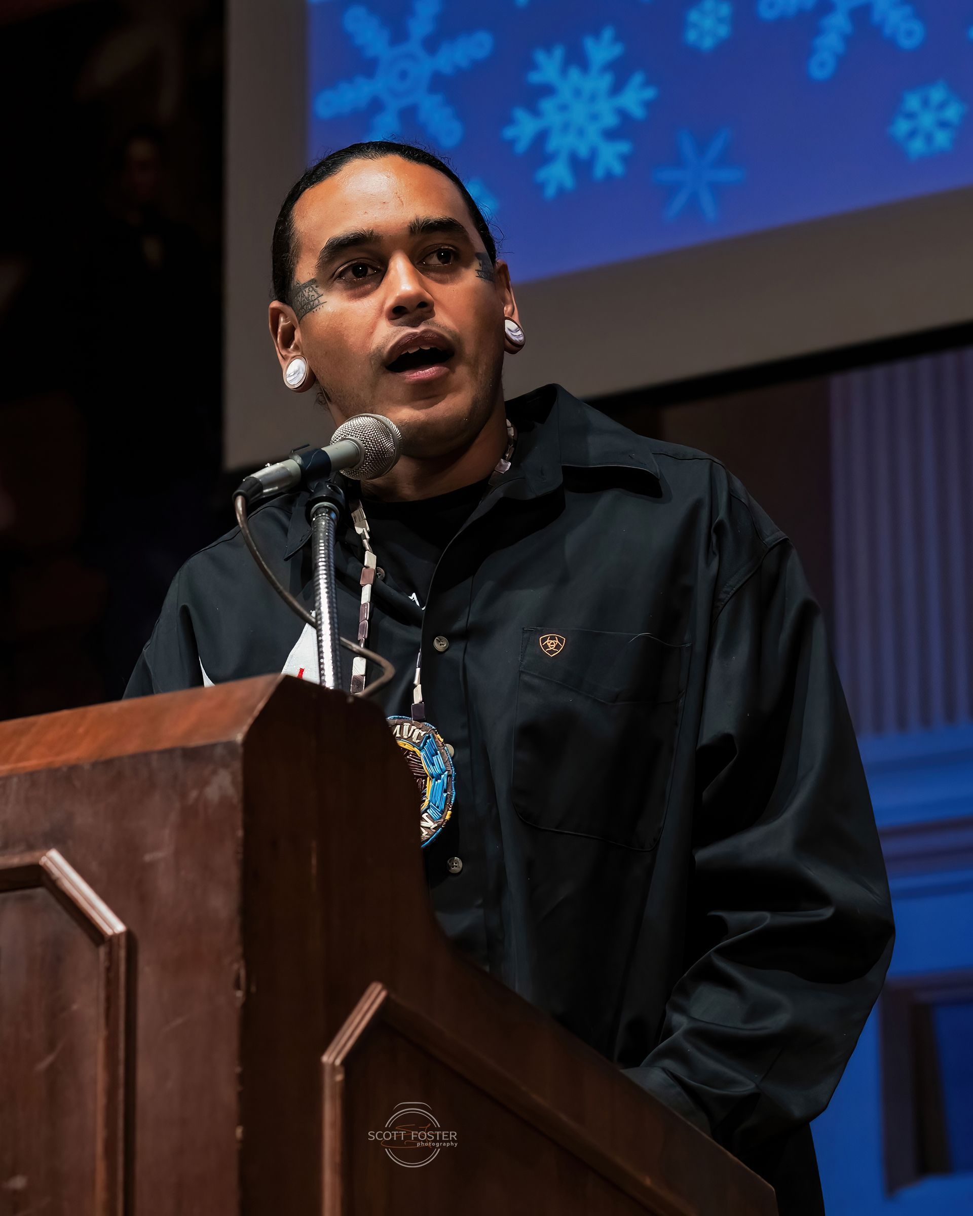Man speaking at a podium, wearing black shirt with ornate jewelry; snowflakes projected behind.