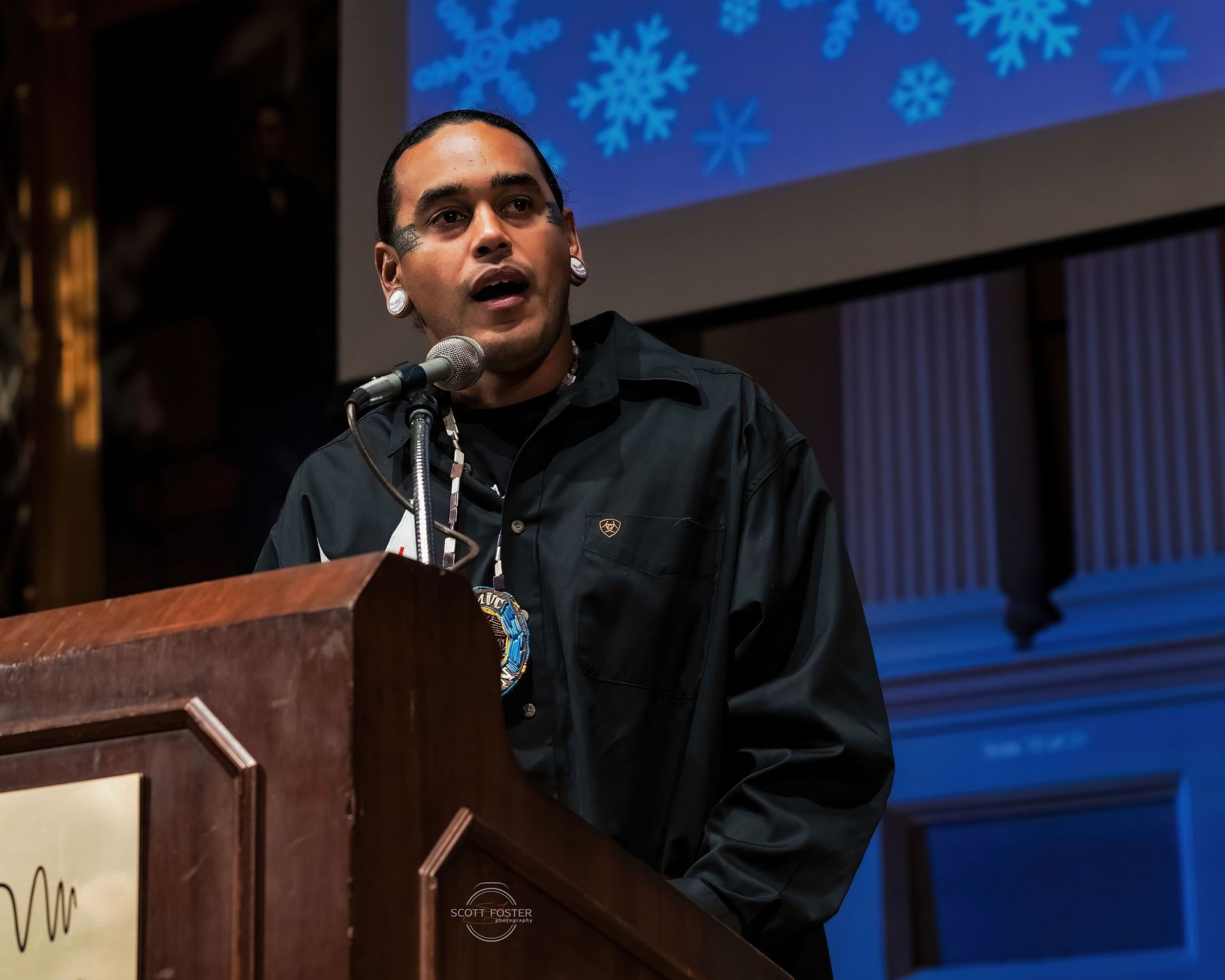 Man speaking at a podium, wearing a dark jacket, in a hall with a snowflake background.