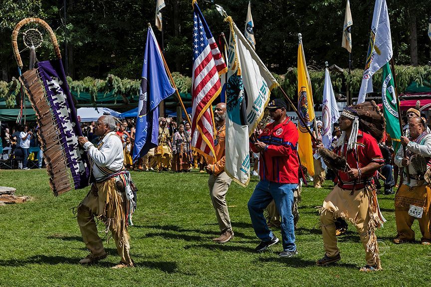 Native Americans carrying flags in a procession on a grassy field; other people watch in the background.