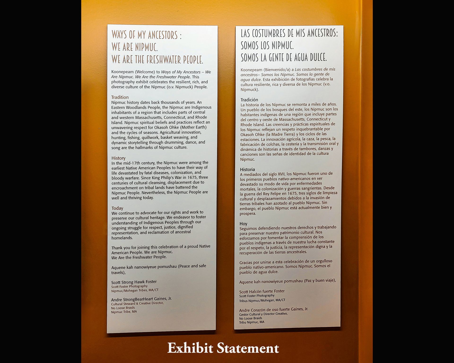 Two white rectangular signs with black text, exhibit statement on an orange wall.