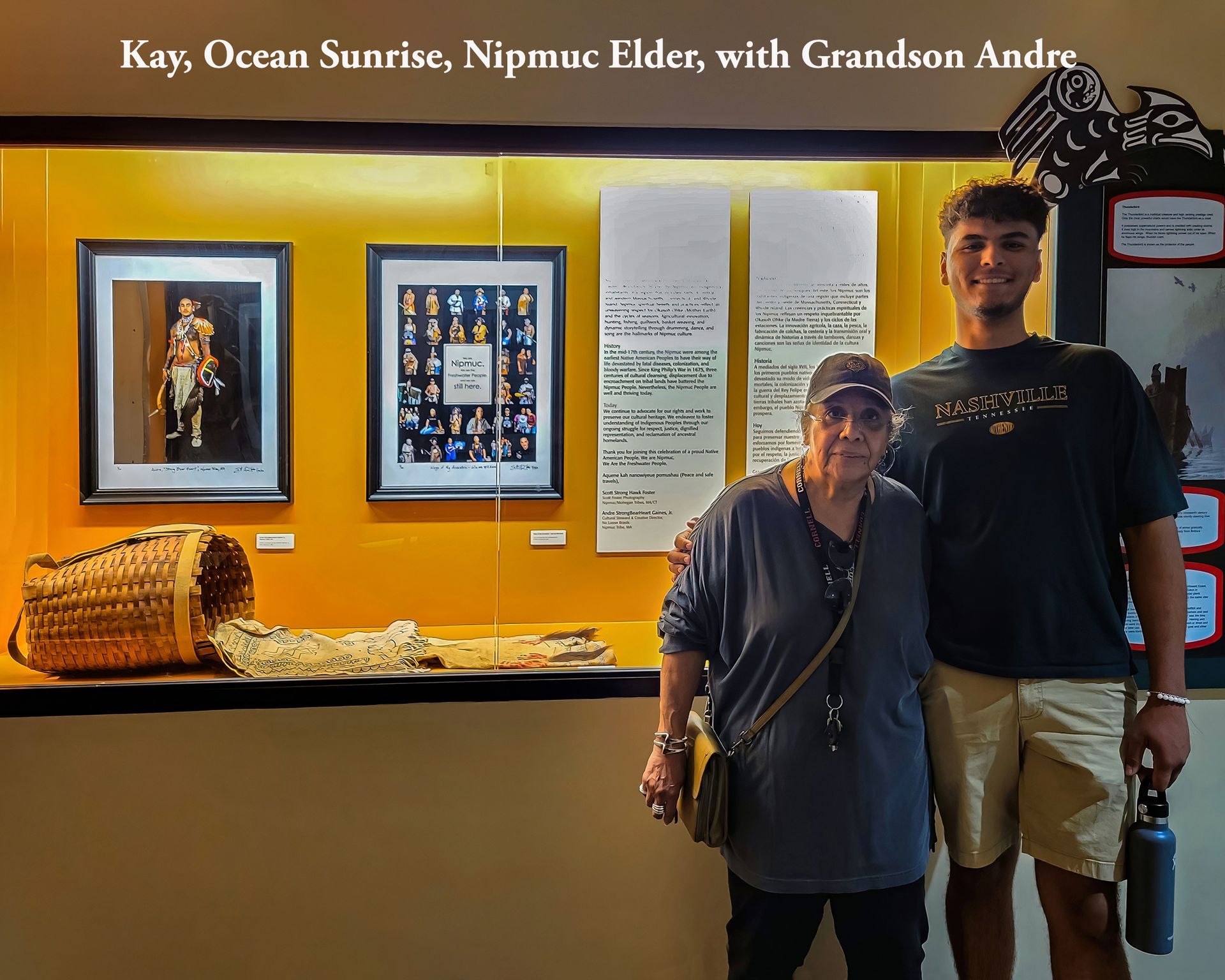 Kay, Nipmuc Elder, and grandson Andre, pose in front of a museum display with baskets and framed photos.