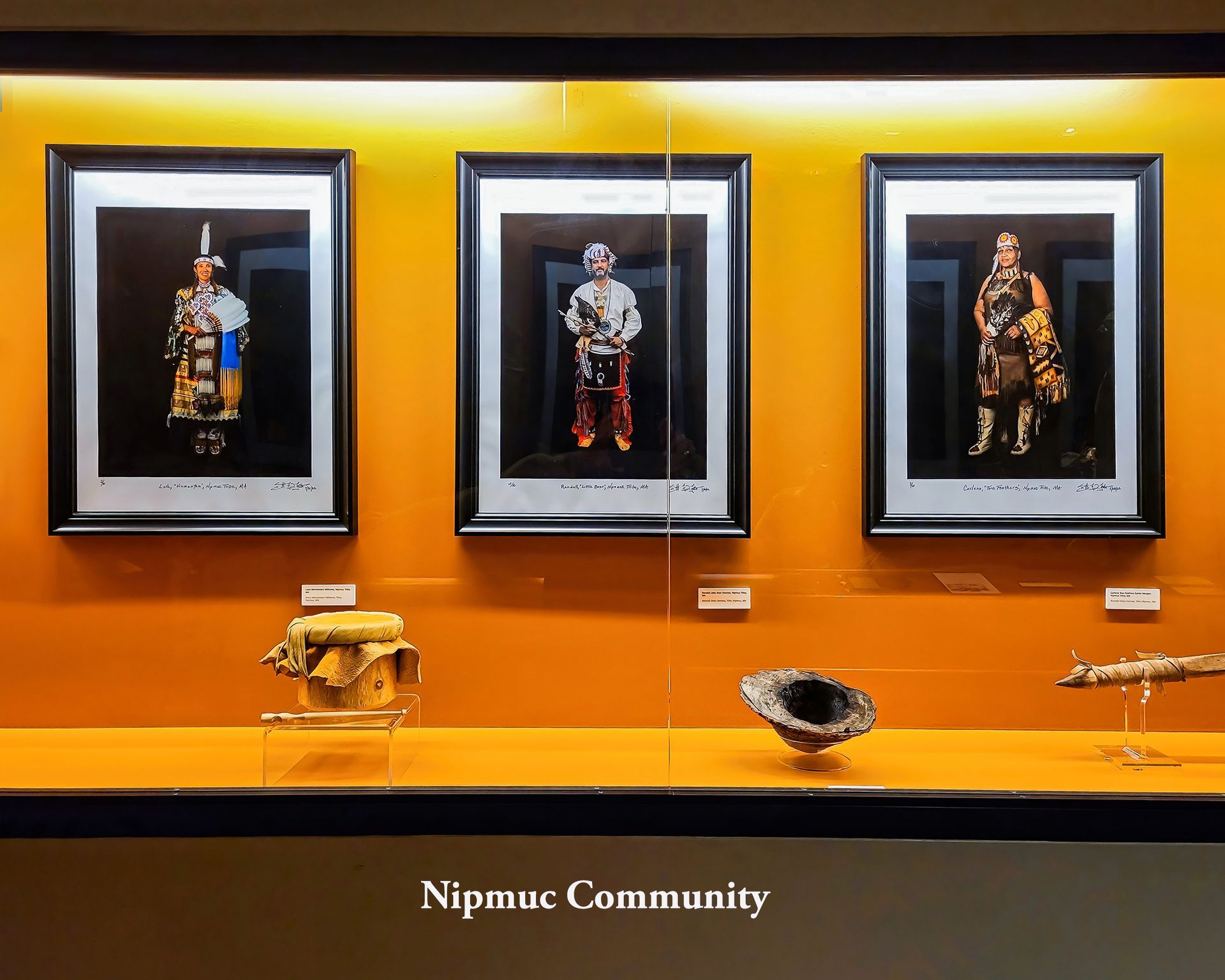 Display case with three framed portraits above artifacts; orange background, Nipmuc Community text.