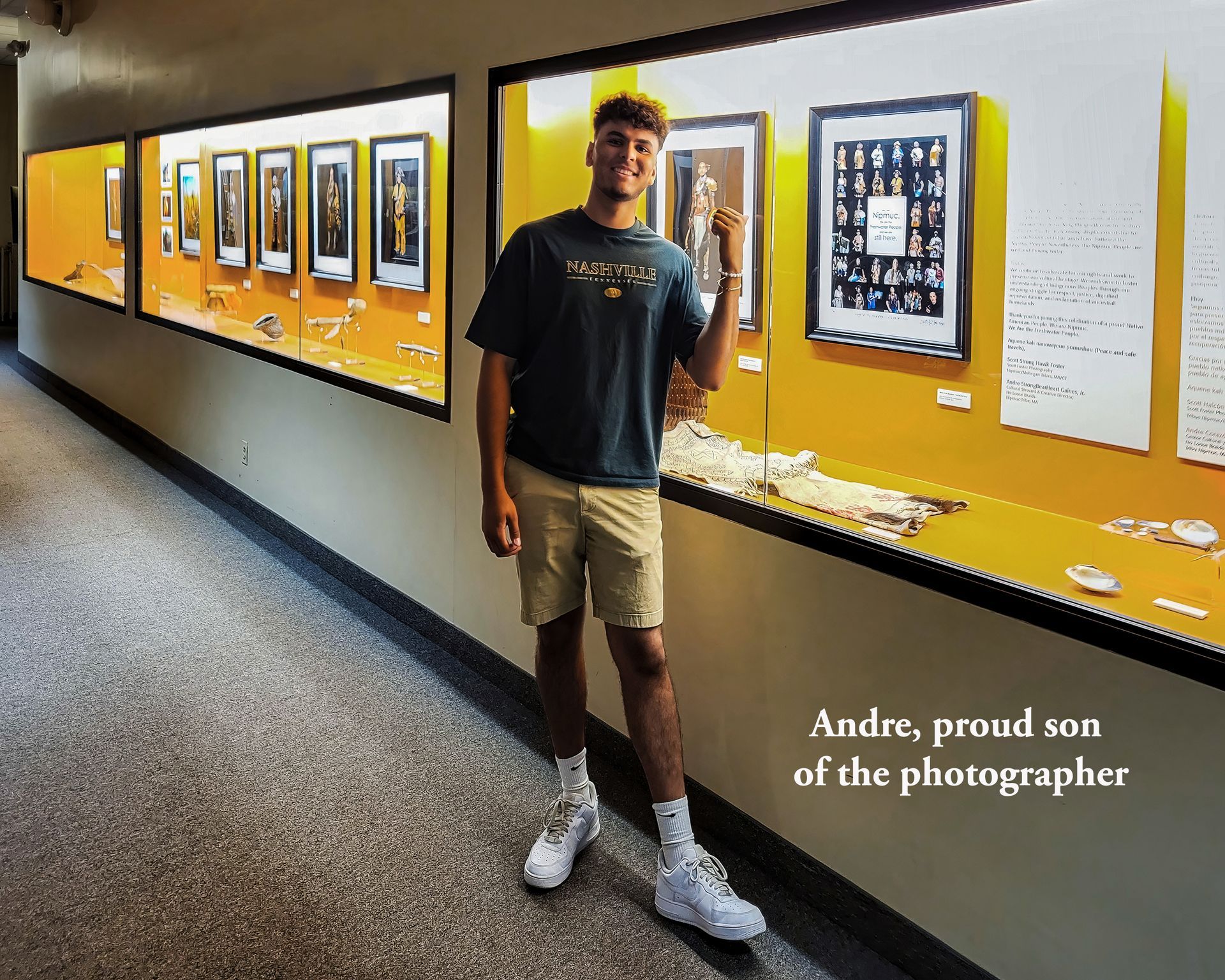 Young man in museum, pointing at a display case. Wall features framed items, yellow background.