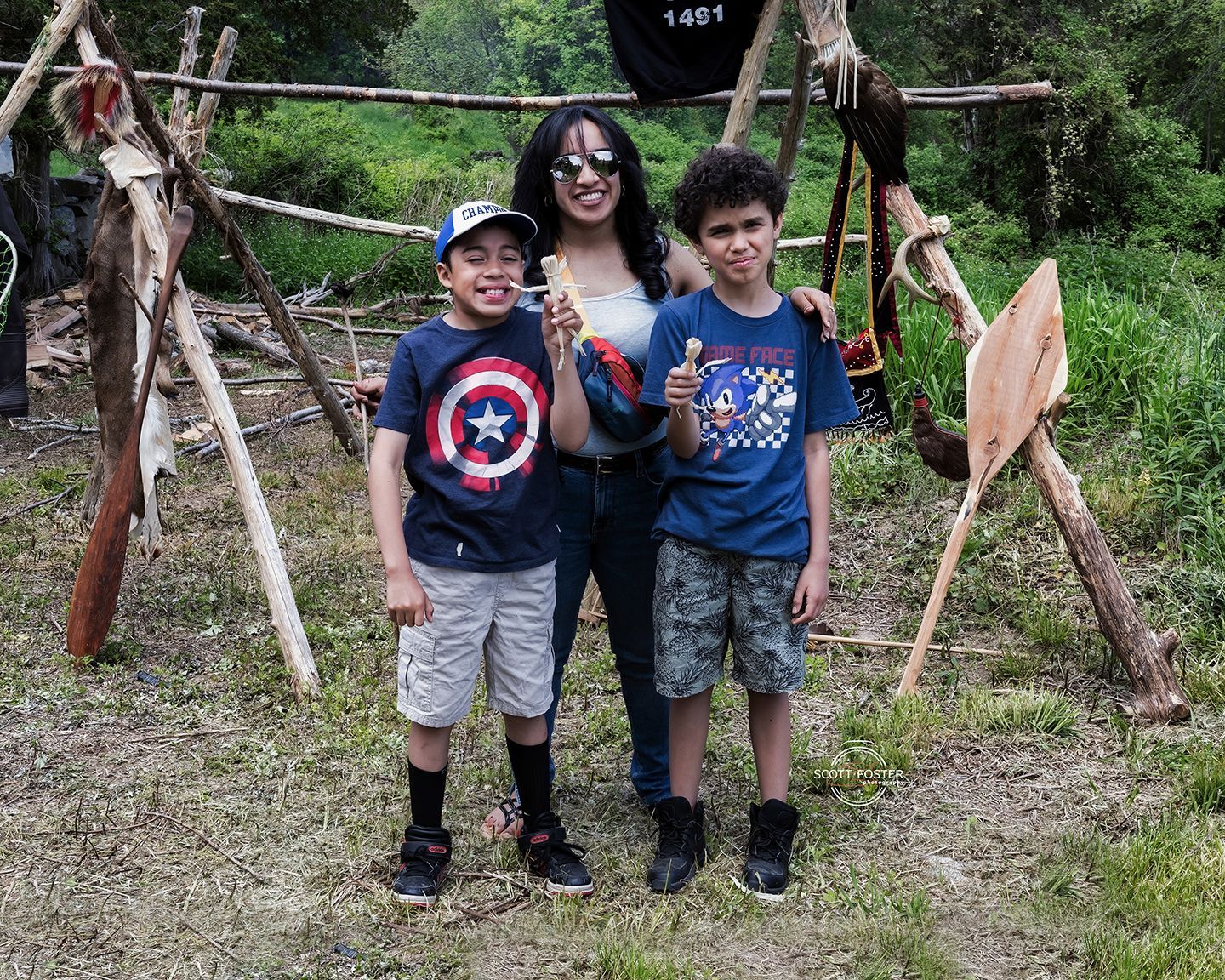 Woman and two boys pose in front of wooden structure, all smiling.