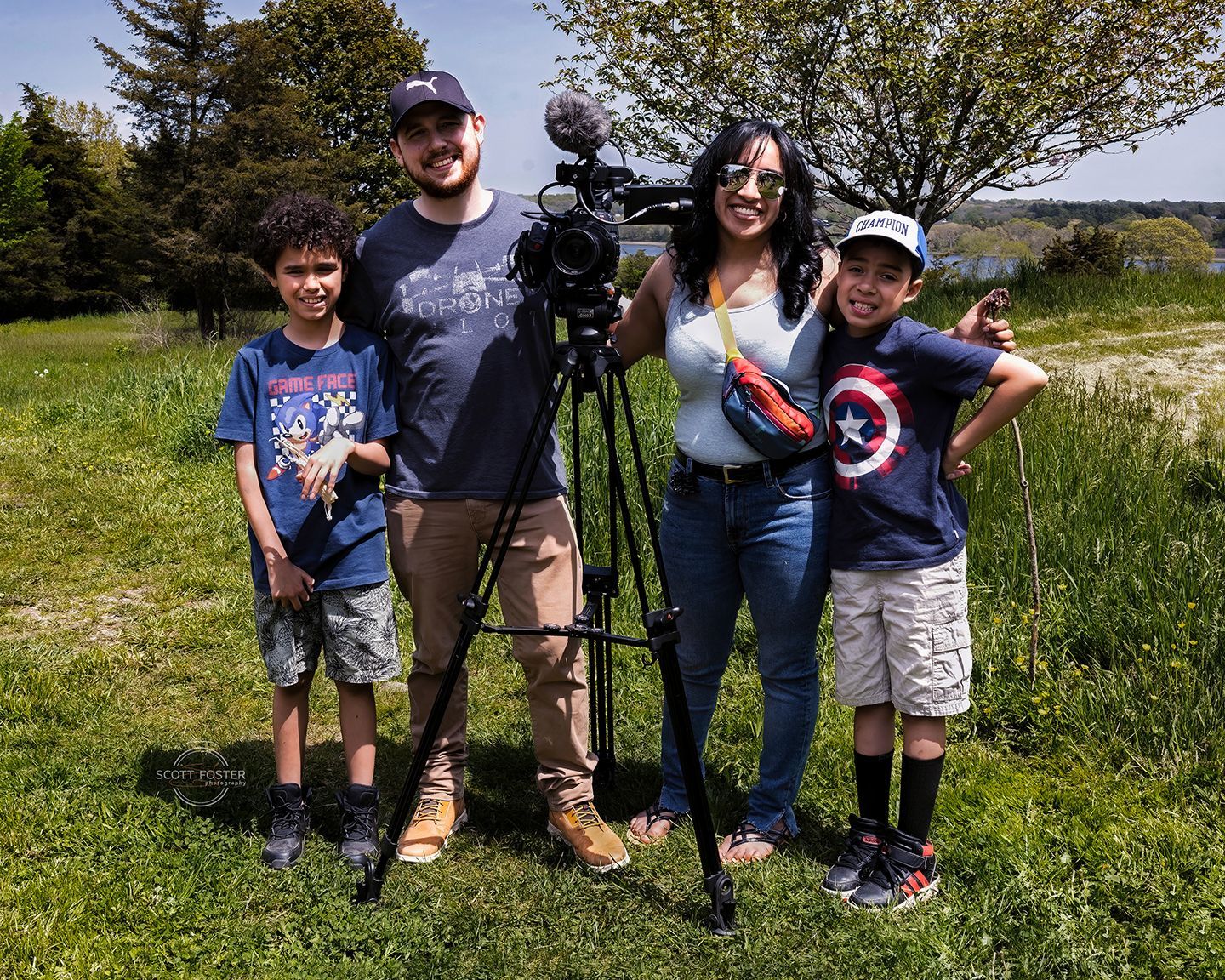Family poses with a video camera on a tripod outdoors, surrounded by green grass.