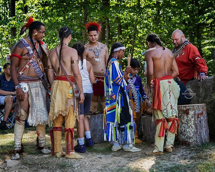 Group of people in Native American clothing, gathered outdoors, some bare-chested, some with face paint.